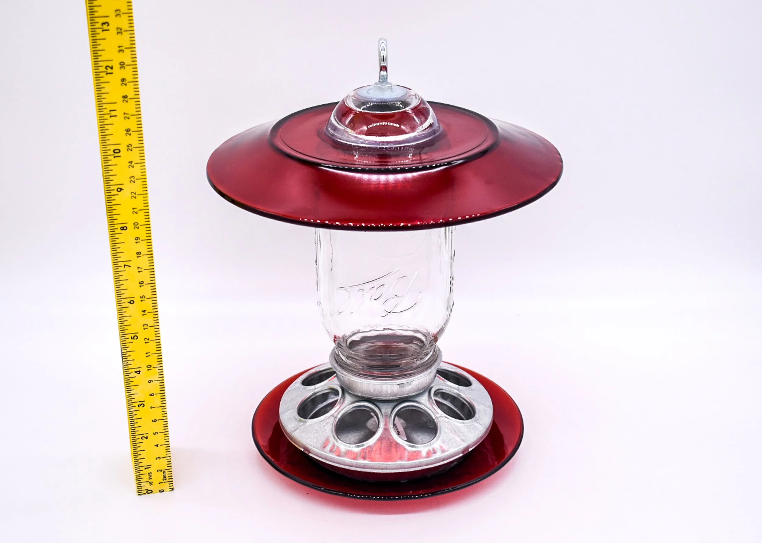 Handmade red glass Mason jar bird feeder with ruler showing size, wide glass rain guard and multiple feeding ports.