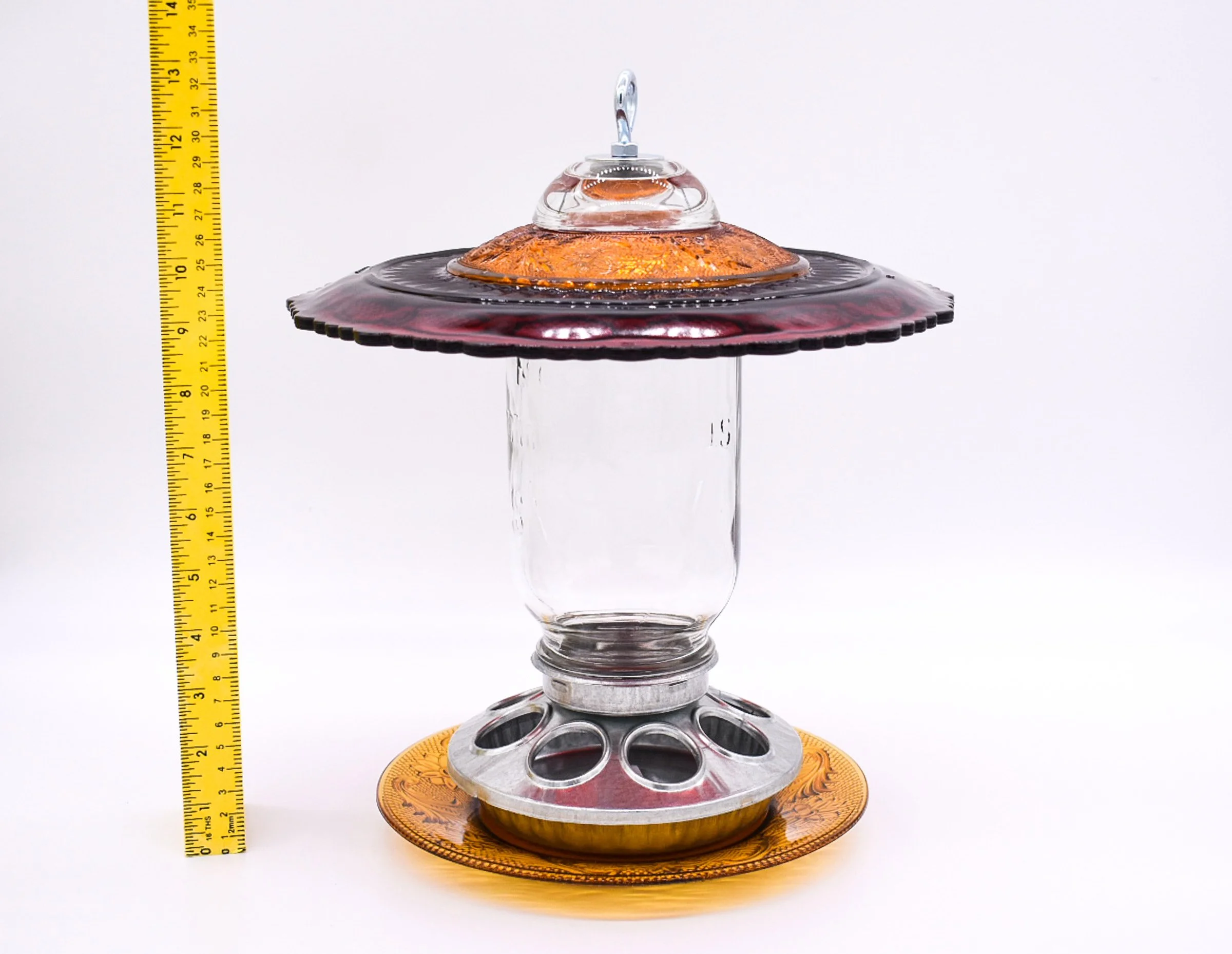 Side view of handmade vintage glass Mason jar seed feeder with ruby red upper plate, amber glass accents, clear quart jar, and measuring tape.