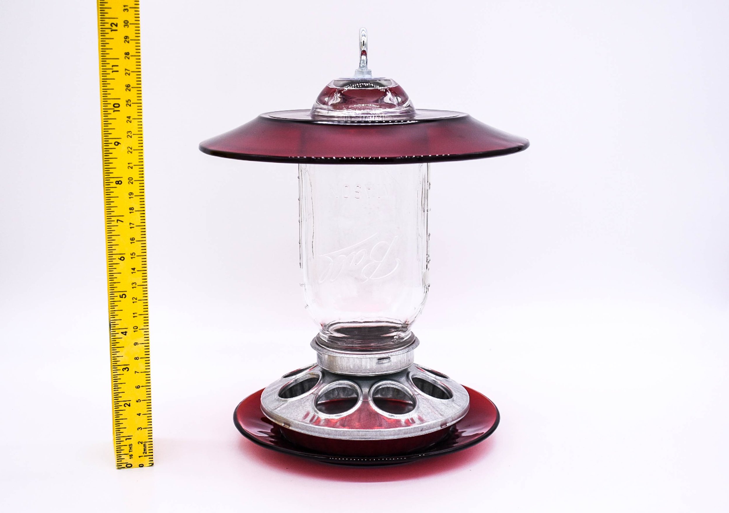 Red glass Mason jar seed bird feeder with ruler showing overall height, glass rain guard and galvanized metal feeding base.