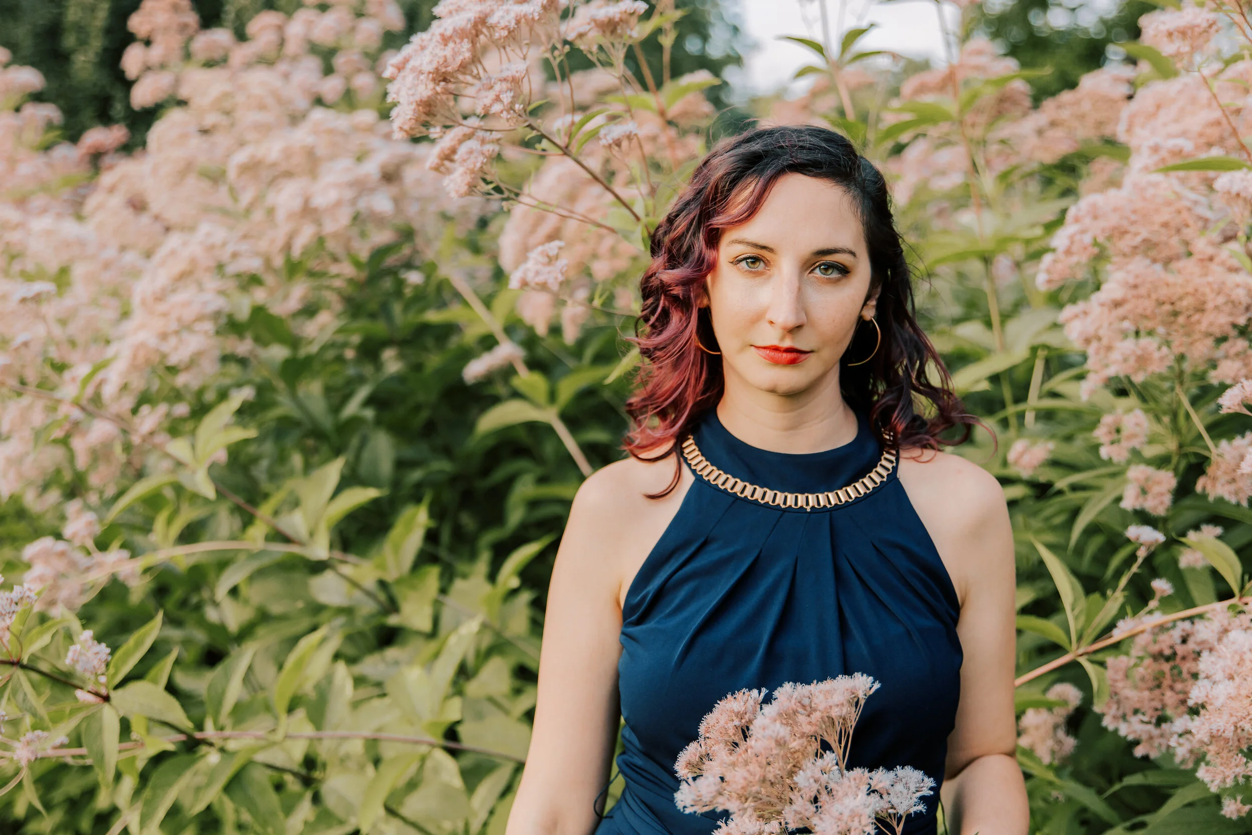 A woman with curly hair dyed with reddish-pink highlights and blue eyes standing outdoors among pink and green flowering bushes, wearing a dark blue sleeveless dress with an embellished neckline.