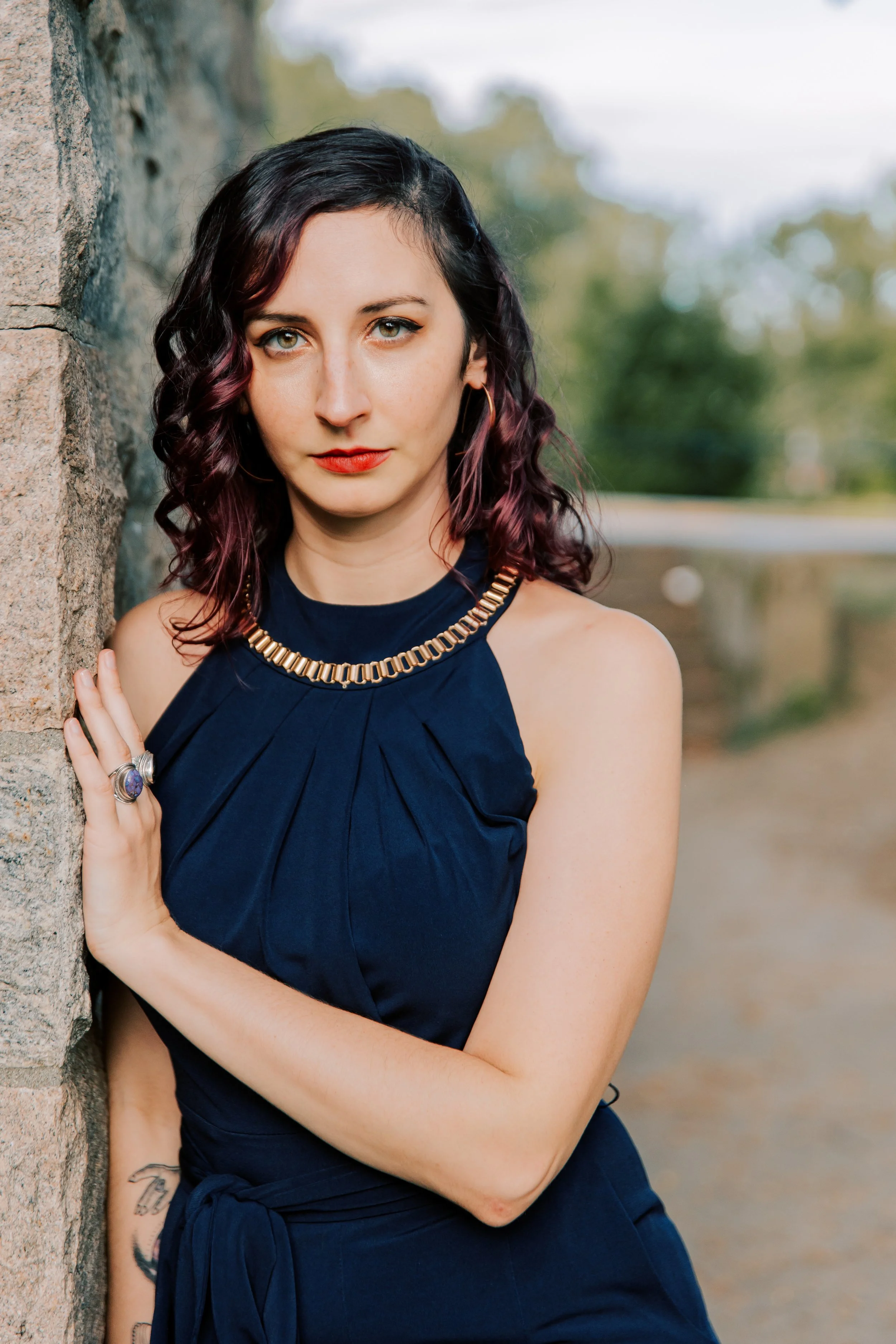 A young woman with dark hair with purple tips, wearing a navy sleeveless dress with a gold necklace, standing outdoors against a brick wall, with a blurred green background.
