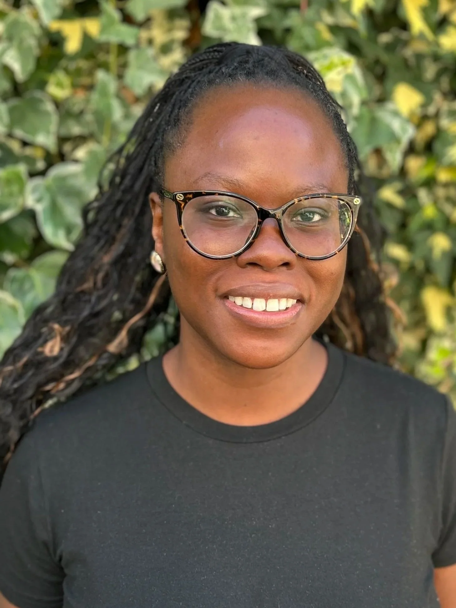 A woman with glasses and long braided hair smiling outdoors in front of green, leafy plants.