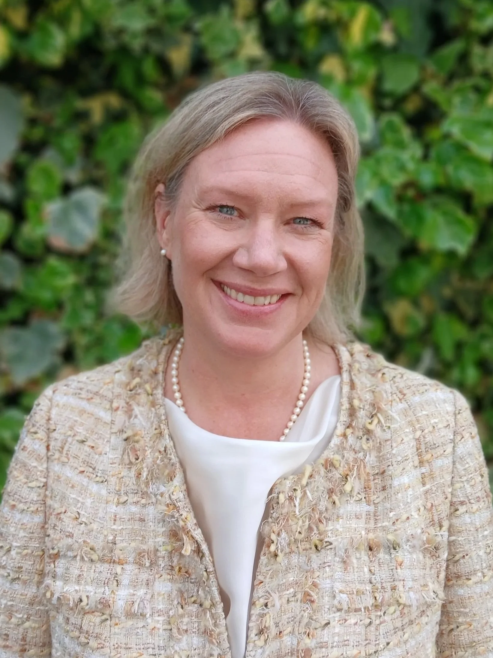 A woman with blonde hair wearing a navy blazer and pearl earrings stands outdoors in front of green leafy bushes.
