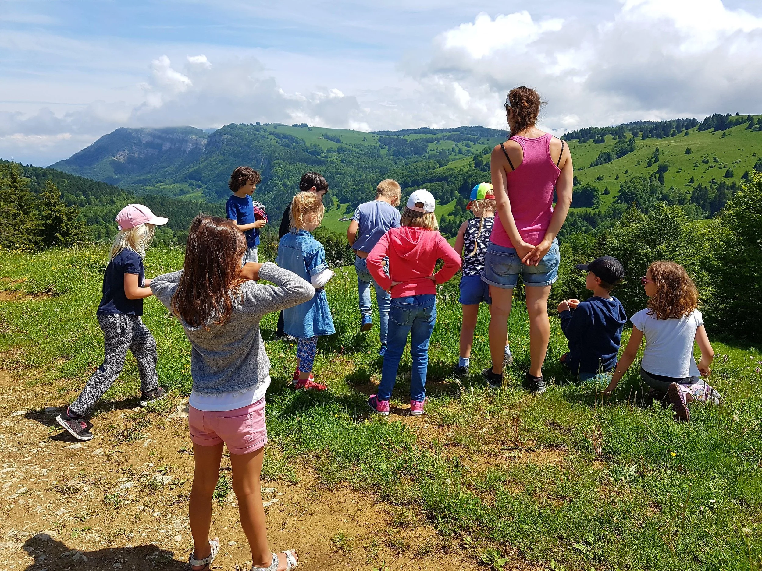 A group of children and an adult woman are on a hike in a lush, green mountain landscape with rolling hills and mountains in the background under a partly cloudy sky.