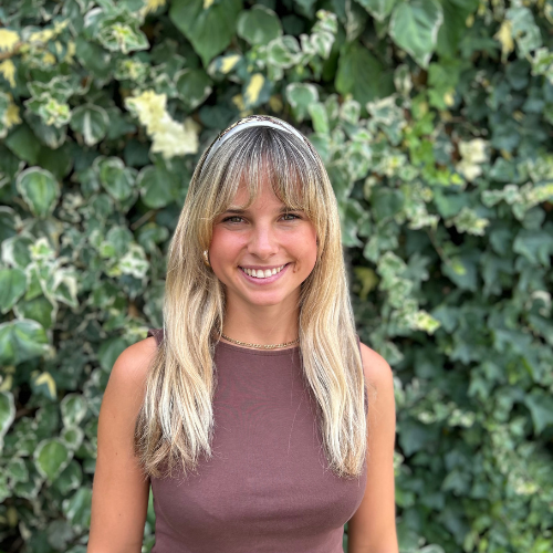 A woman with short light brown hair and blue eyes smiling, wearing a white shirt, standing outdoors with green leafy bushes in the background.