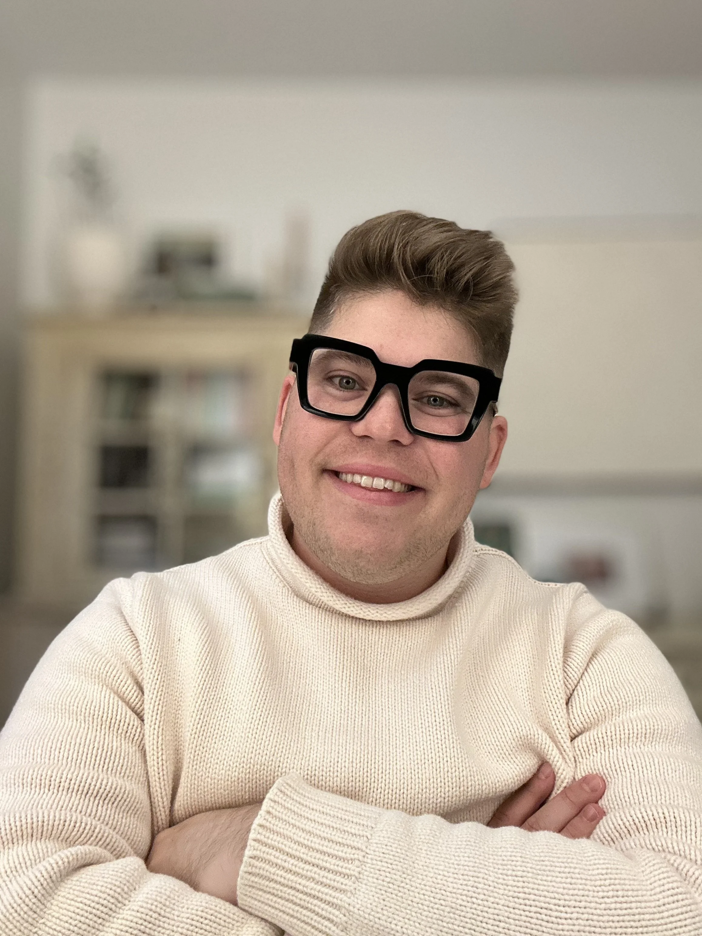 A smiling man wearing large black glasses and a cream-colored turtleneck sweater, crossing his arms, standing in a blurred indoor background.