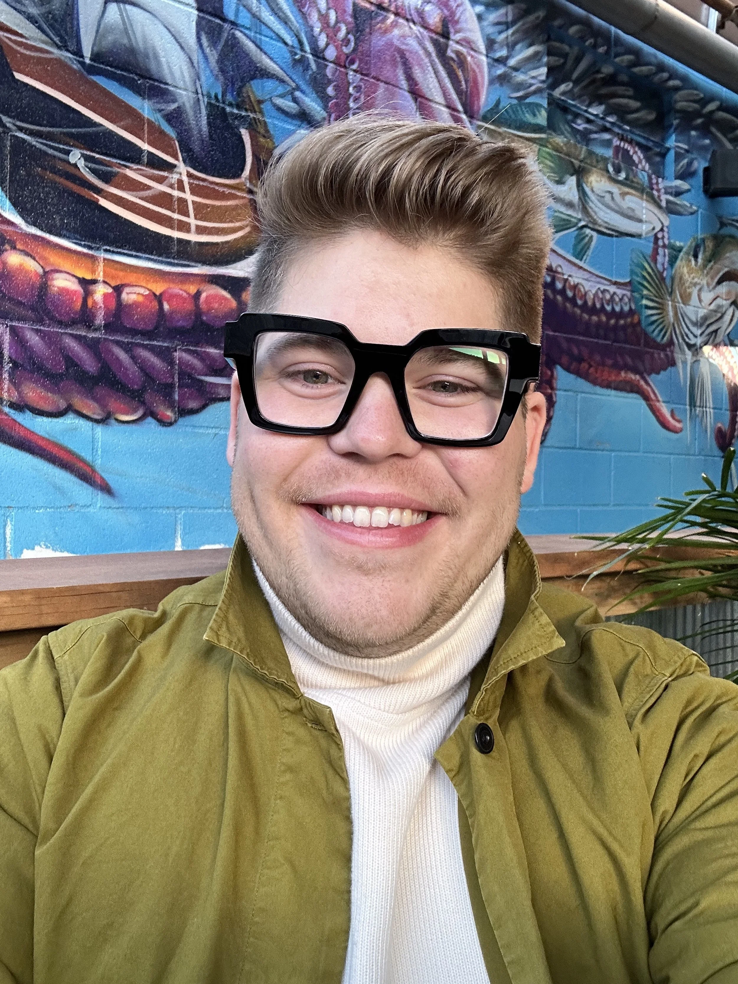 A smiling young man wearing large black glasses, a beige turtleneck, and an olive green jacket, sitting indoors against a colorful painted wall with abstract art and fish imagery.