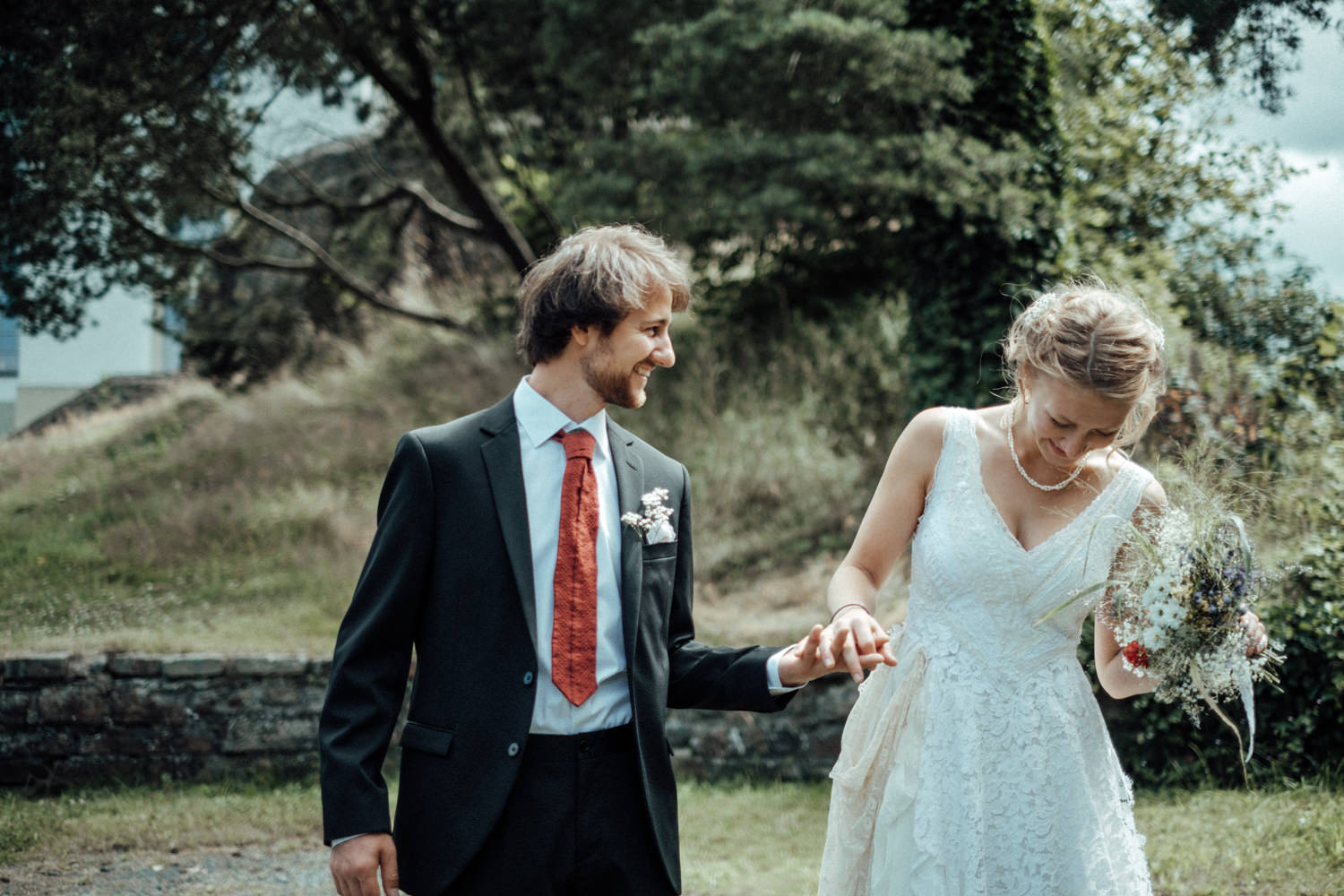 Hochzeitspaar hält Händchen und lacht glücklich in Natur