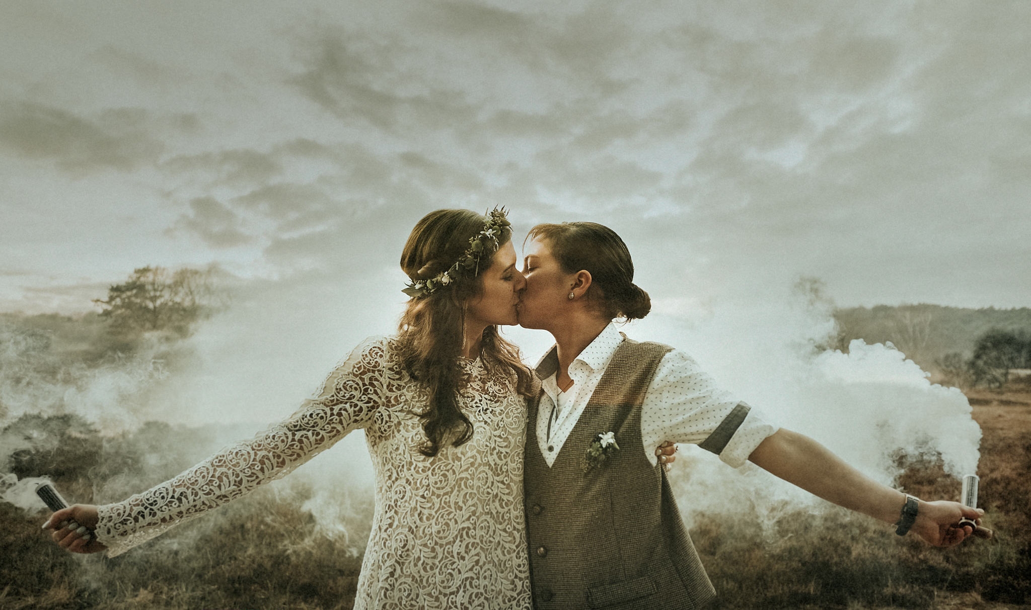 Alternatives gleichgeschlechtliches Hochzeitspaar sich küssend beim Paarshooting mit Rauchfackeln in der Natur.