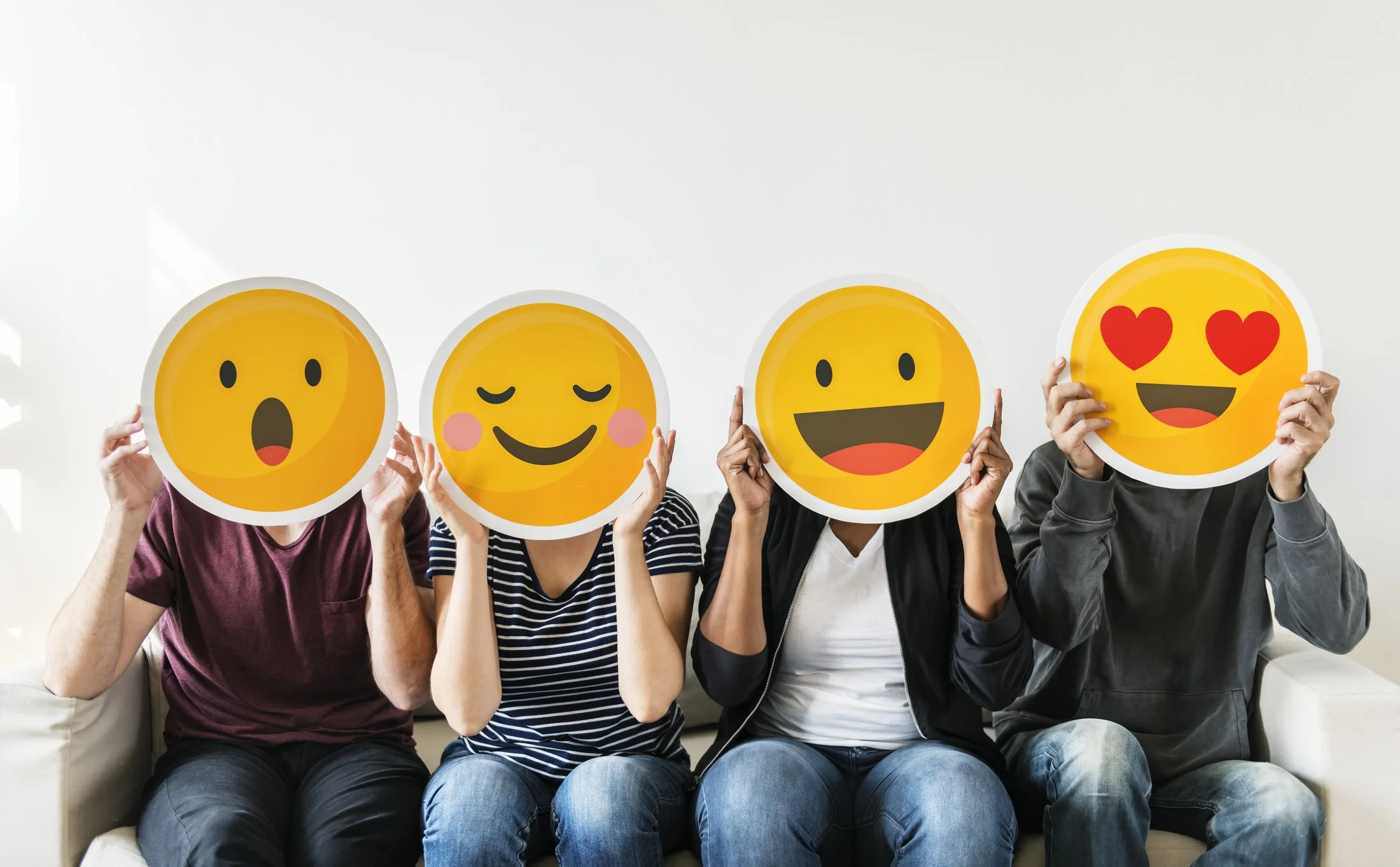 Four people sitting on a couch holding emoji face masks in front of their faces, with each mask displaying a different emoji expression.