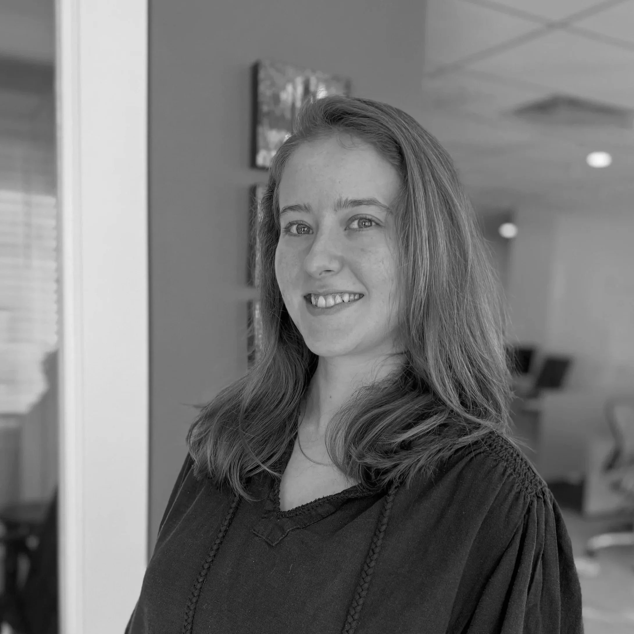 A woman with long wavy hair smiling, wearing a black top, in an indoor setting with paintings on the wall in the background.