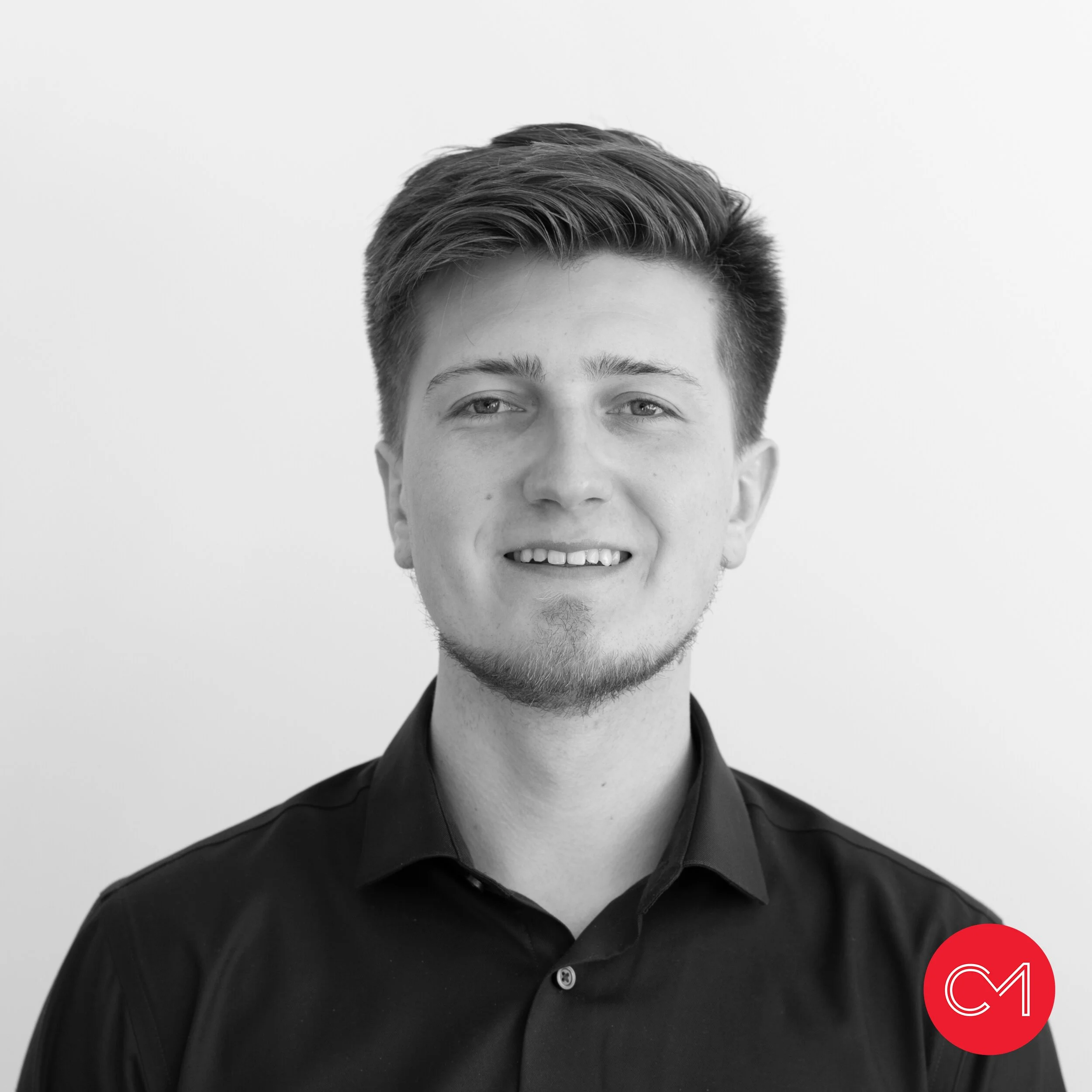 Black and white portrait of a young man with short, styled hair, light facial hair, and a slight smile, wearing a dark button-up shirt, against a plain background with a red circular logo containing white stylized 'CM' in the bottom right corner.