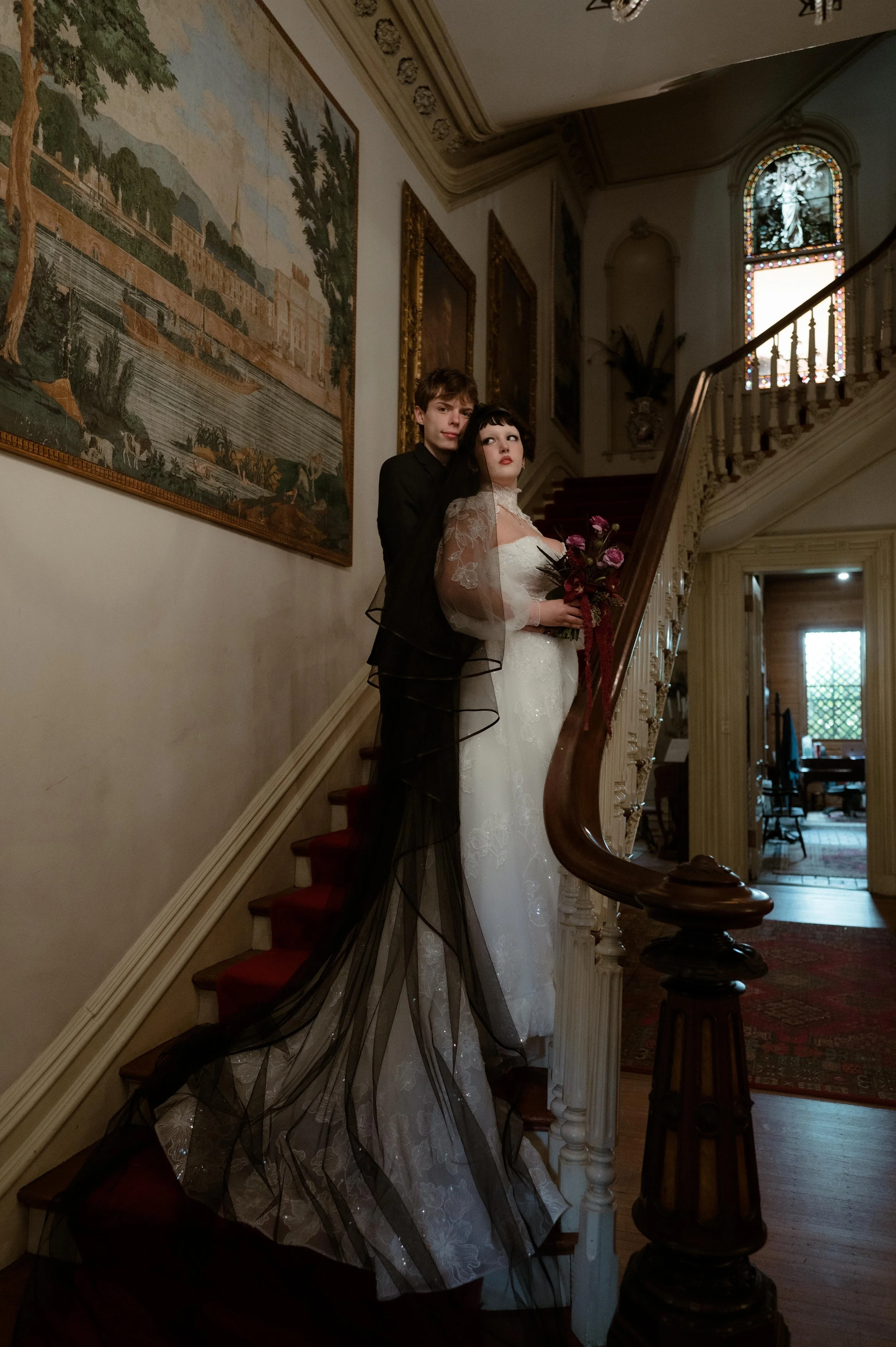 A bride and groom standing on the staircase in the foyer of the Woodruff-Fontaine house in Memphis, Tennessee. The bride wears a sparkly white wedding gown with attached sleeves and high neckline. She has short black hair and wears a long black veil.