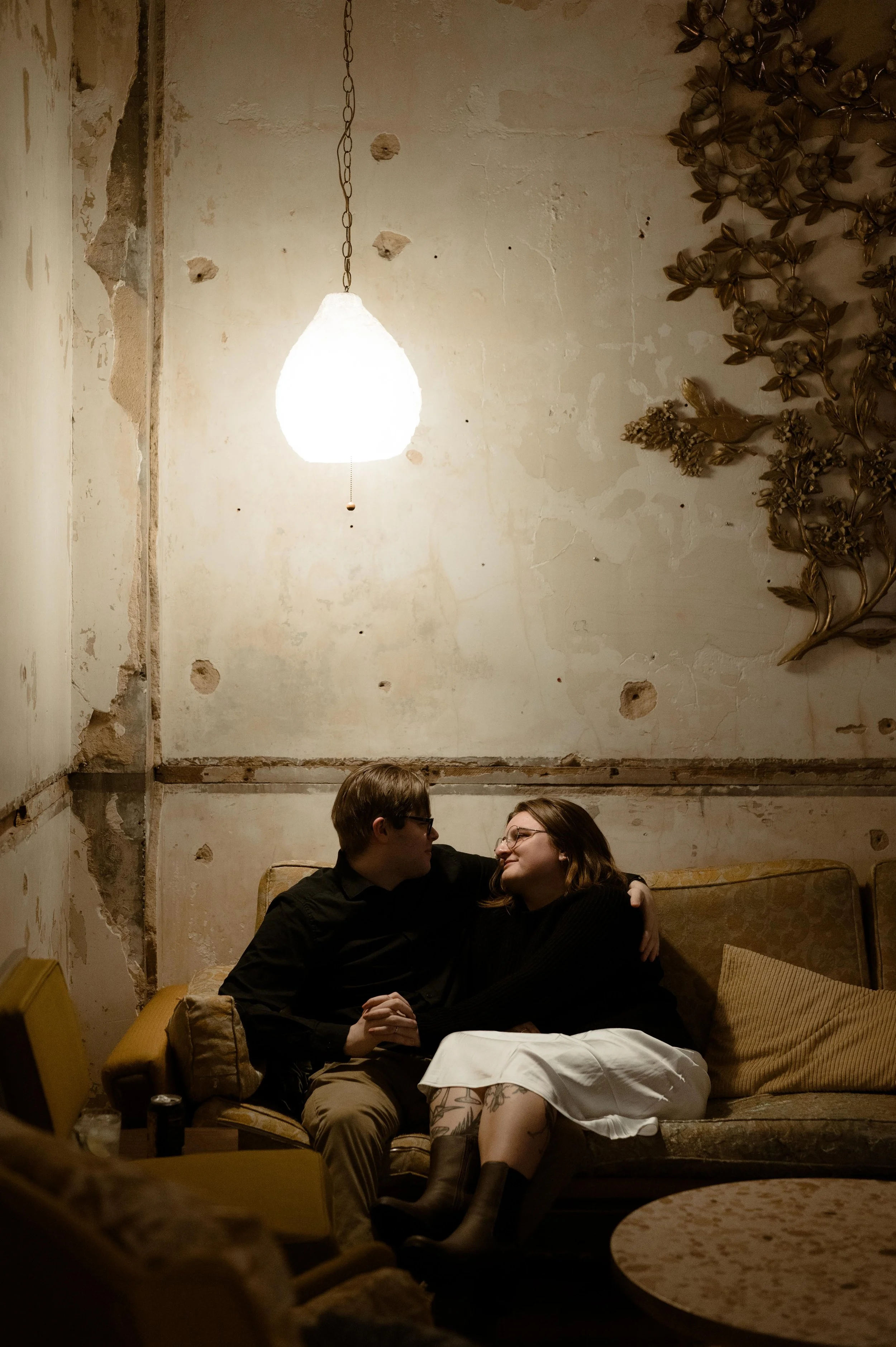 A couple snuggles on a couch for their engagement session at Art Bar at Crosstown Concourse in Memphis, Tennessee. The lighting is moody, overhead light from a hanging lamp. They both wear black and brown, and one wears white skirt with boots.