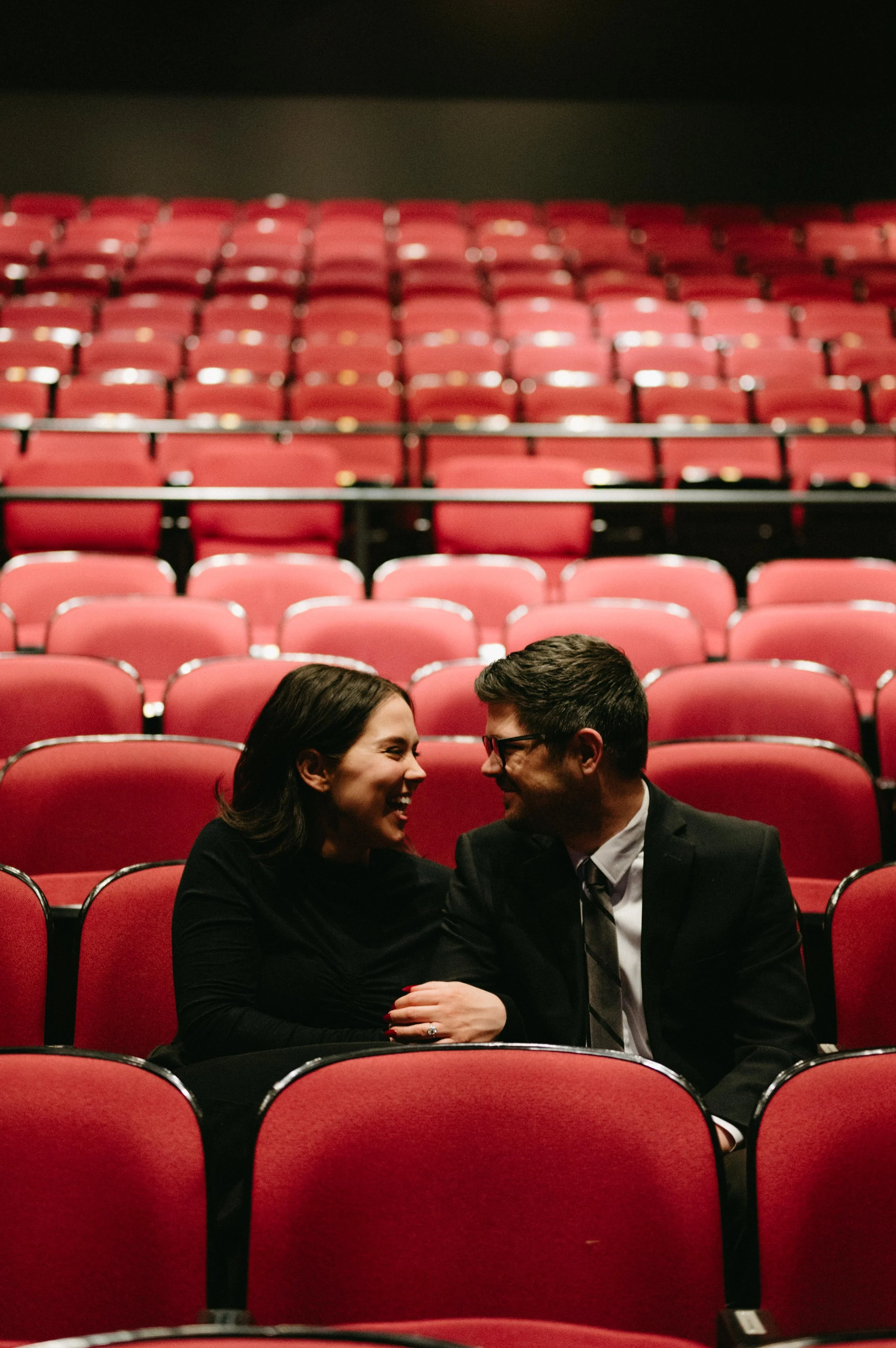 A couple sits in the audience of Theater Memphis in Memphis, Tennessee for their engagement photos. The seats are red, they are wearing all black, and looking at each other and smiling.
