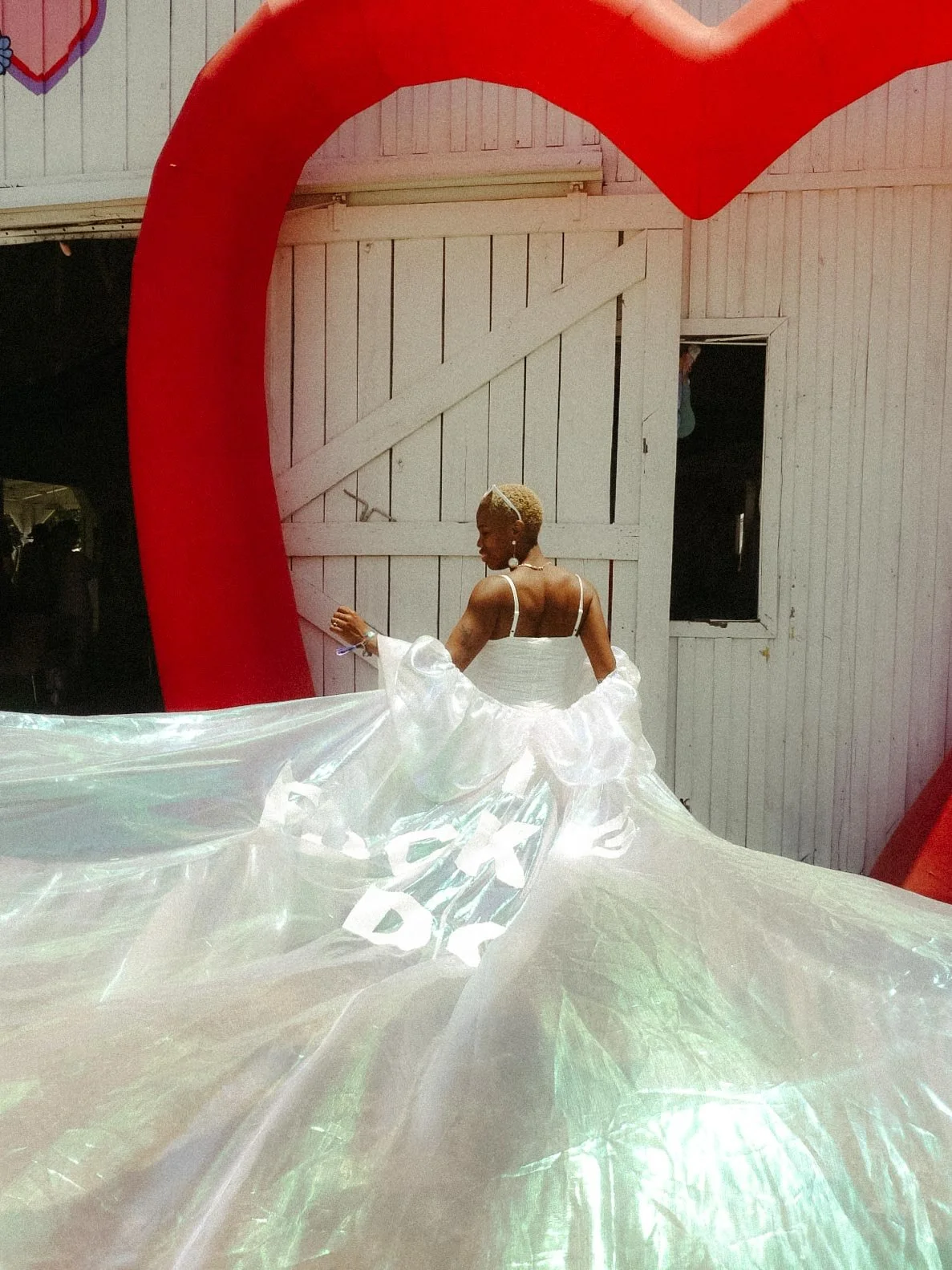 A bride posing outside of the marriage chapel at Bonnaroo outside Nashville, Tennessee. She wears a white sleeveless dress and a long white cape that shimmers green and blue. The Bonnaroo marriage chapel is white with a big red blow up heart.