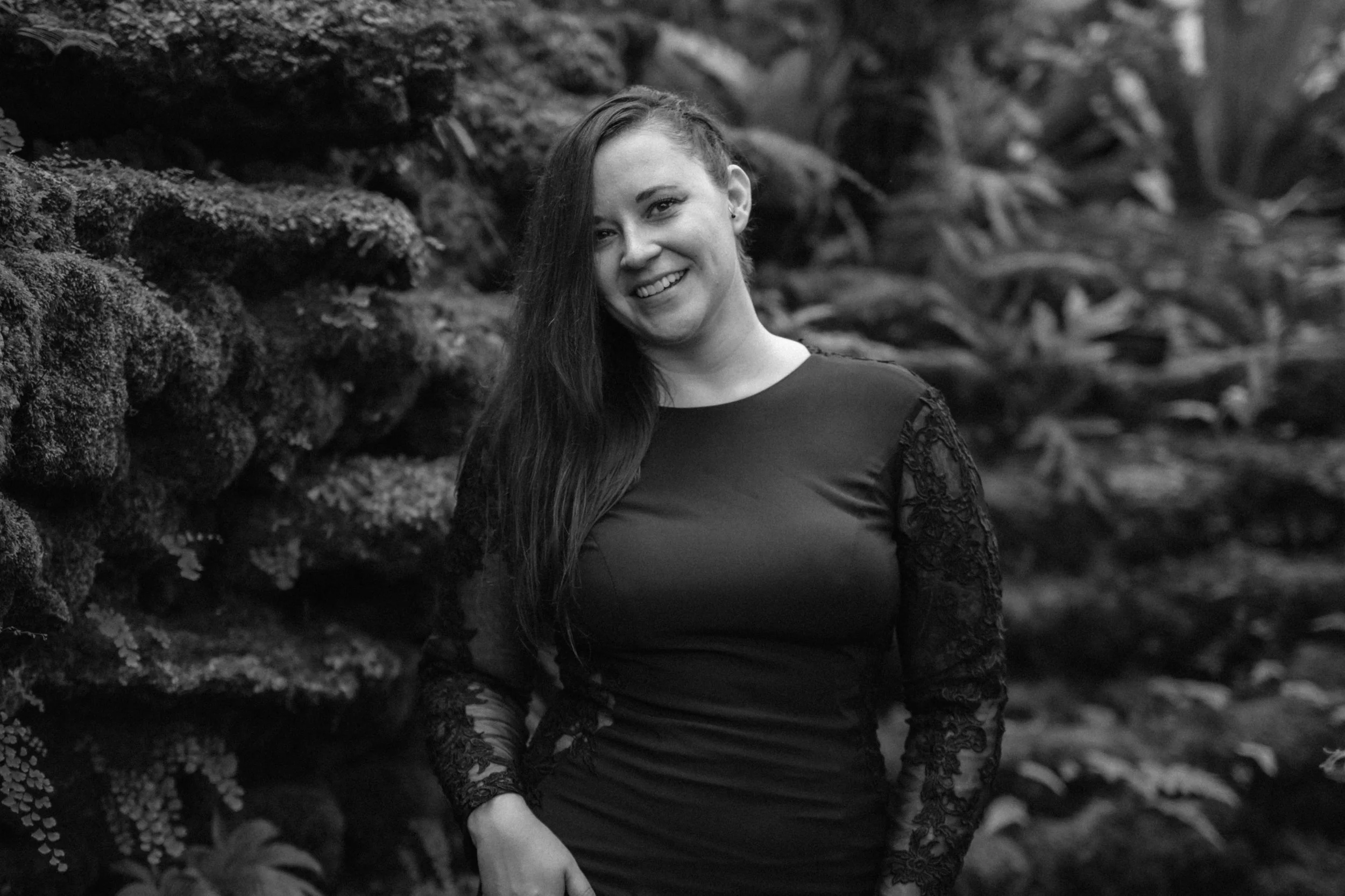 Black and white portrait of a bride in an all black gown with long black lace sleeves and her hair slicked back to one side. She is smiling at the camera.