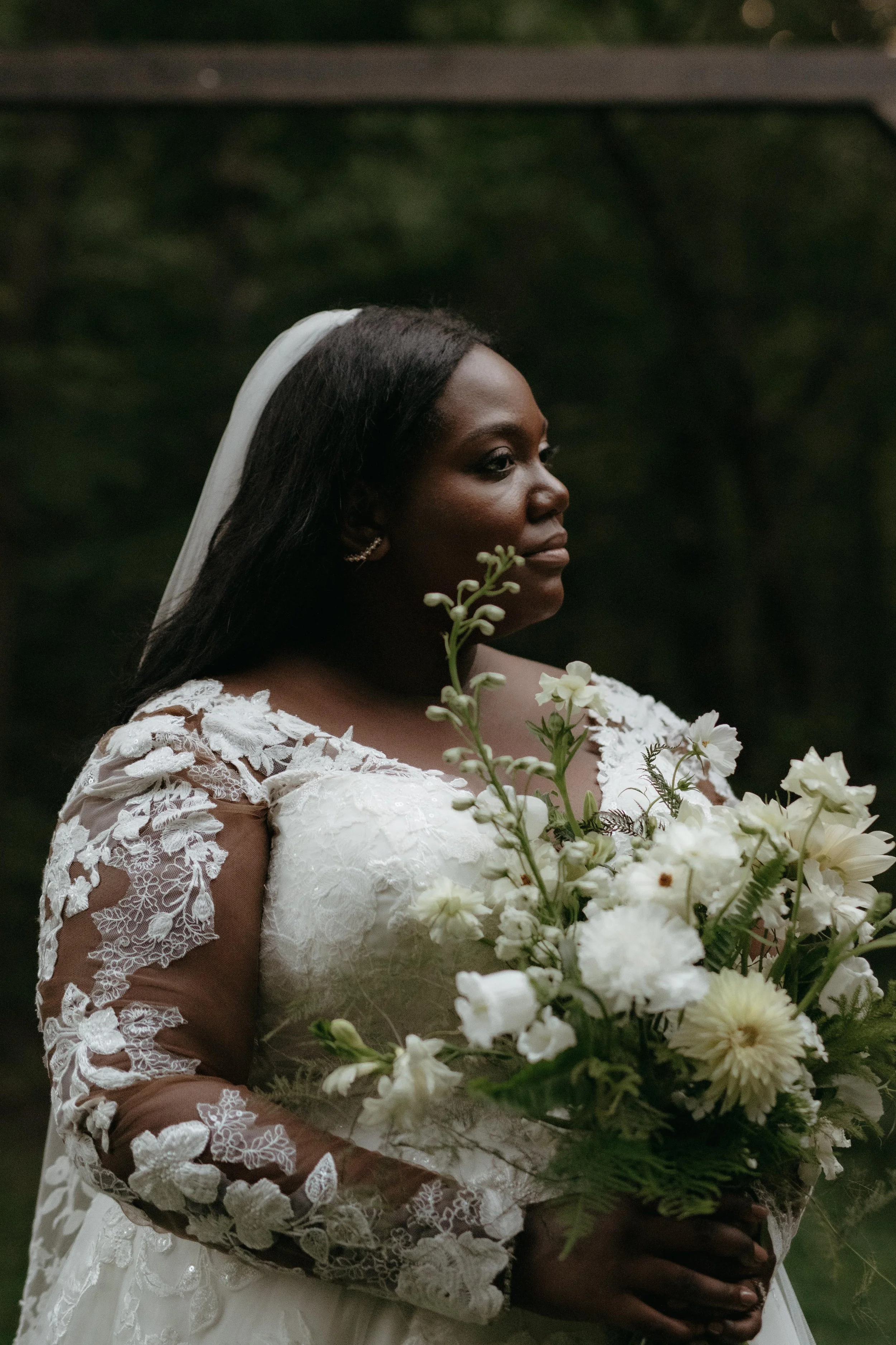 Bridal portrait taken at Lichterman Nature Center in Memphis, Tennessee. The bride holds a bouquet of white flowers and greenery, and wears a white wedding dress with lace, and long sheer lace sleeves with flower embroidery. She wears a simple veil.
