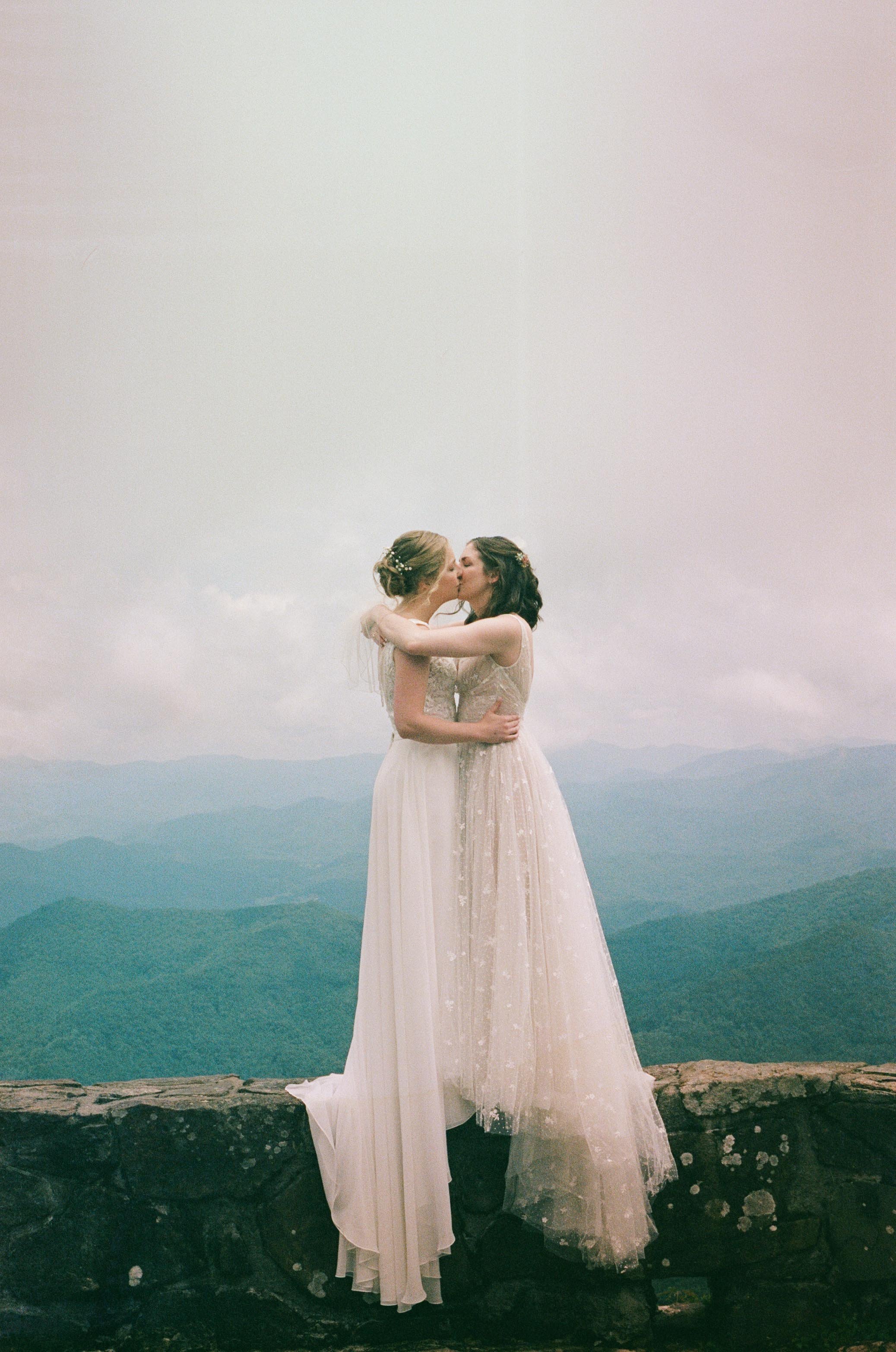 Two brides kiss and embrace on the Wayah Bald outlook in Nantahala, North Carolina. Behind them is a cloudy grey sky and green and blue hills. One bride wears an off white dress with small embroidered flowers and tulle.
