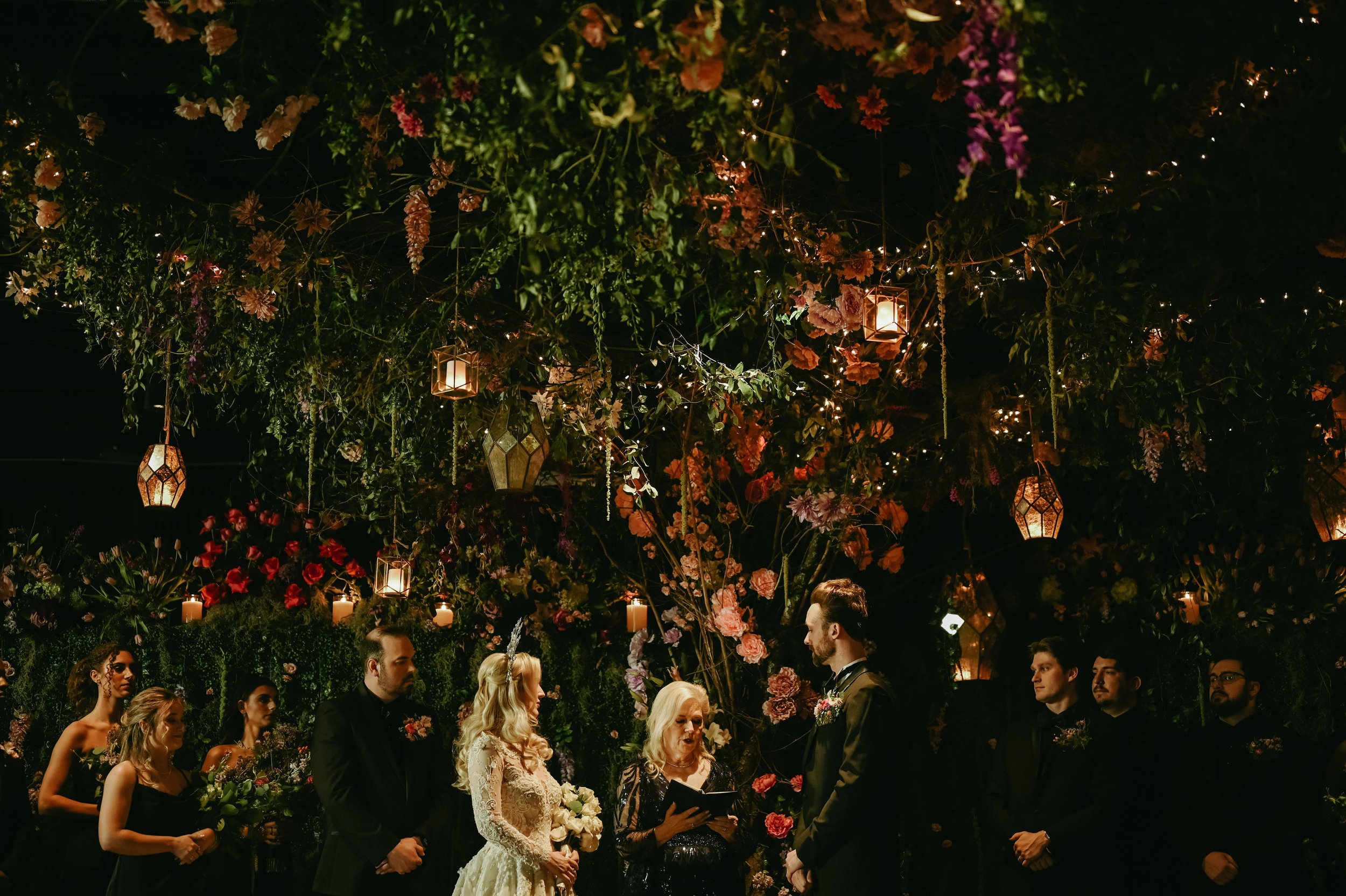 A marriage ceremony at the Kent in Memphis, Tennessee under a floor to ceiling cover of green, red, pink, and orange flowers created by Lynn Doyle Flowers, the bride wears her long blonde hair down and has a crown on top of her head, and she holds an