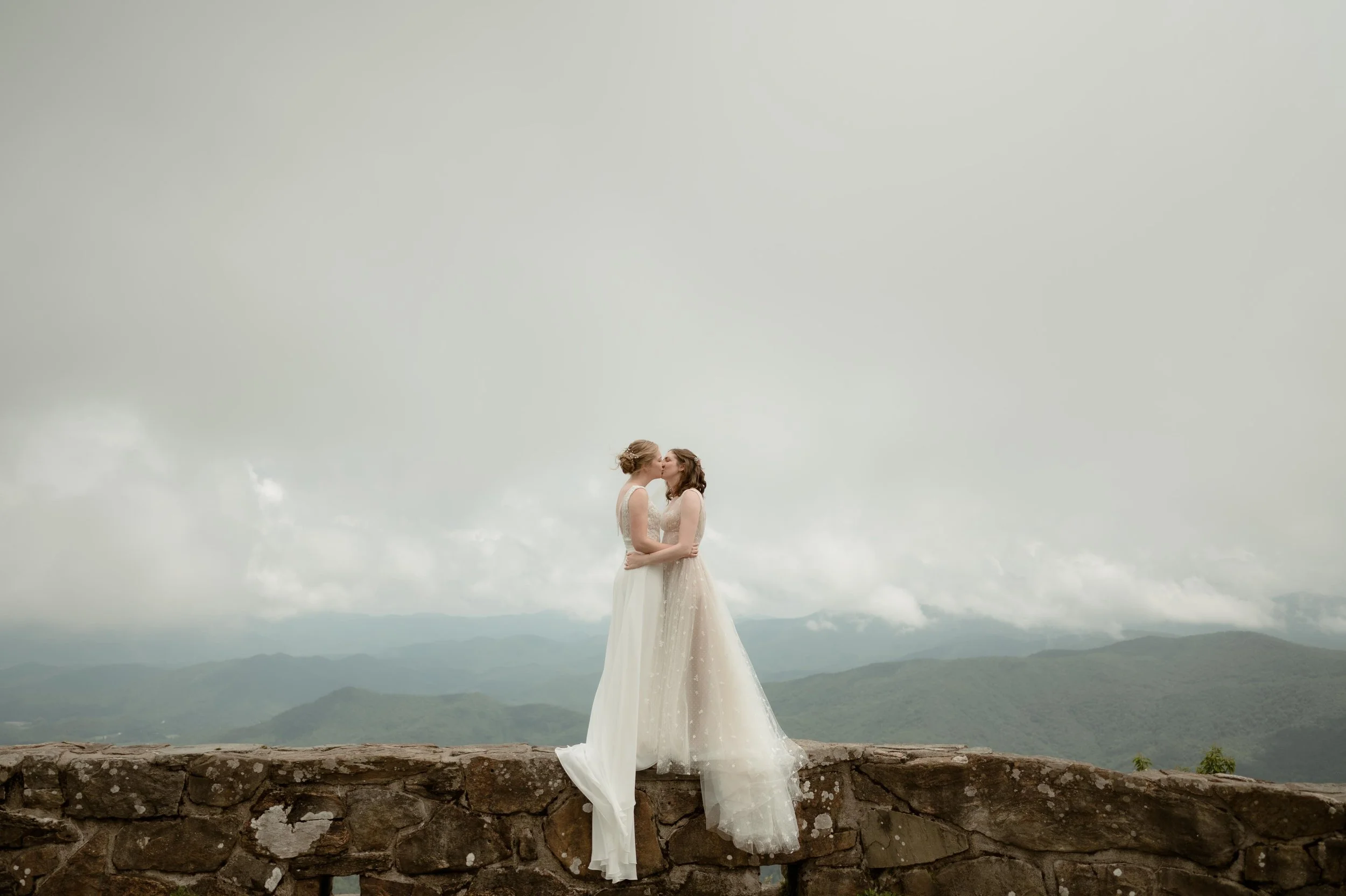 Moody North Carolina wedding photographer two brides kiss on top of Wayah Bald in Nantahala. North Carolina with a view of the Appalachian mountains behind them. Moody Memphis wedding photographer. Documentary North Carolina wedding photographer.