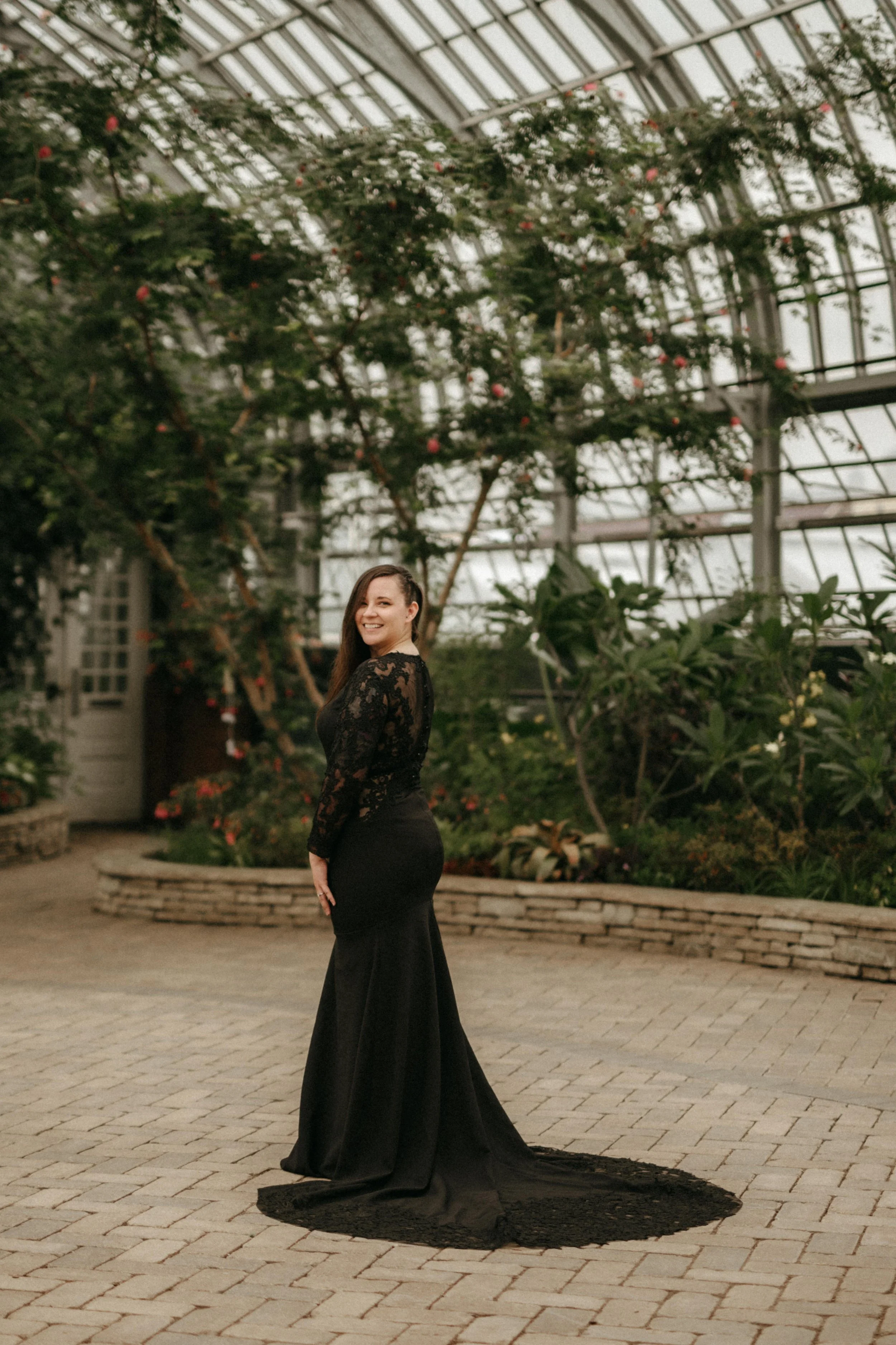 A bride in an all black wedding dress with long black lace sleeves looks over her shoulder at the camera. She stands in the Garfield Park Conservatory in Chicago, Illinois.