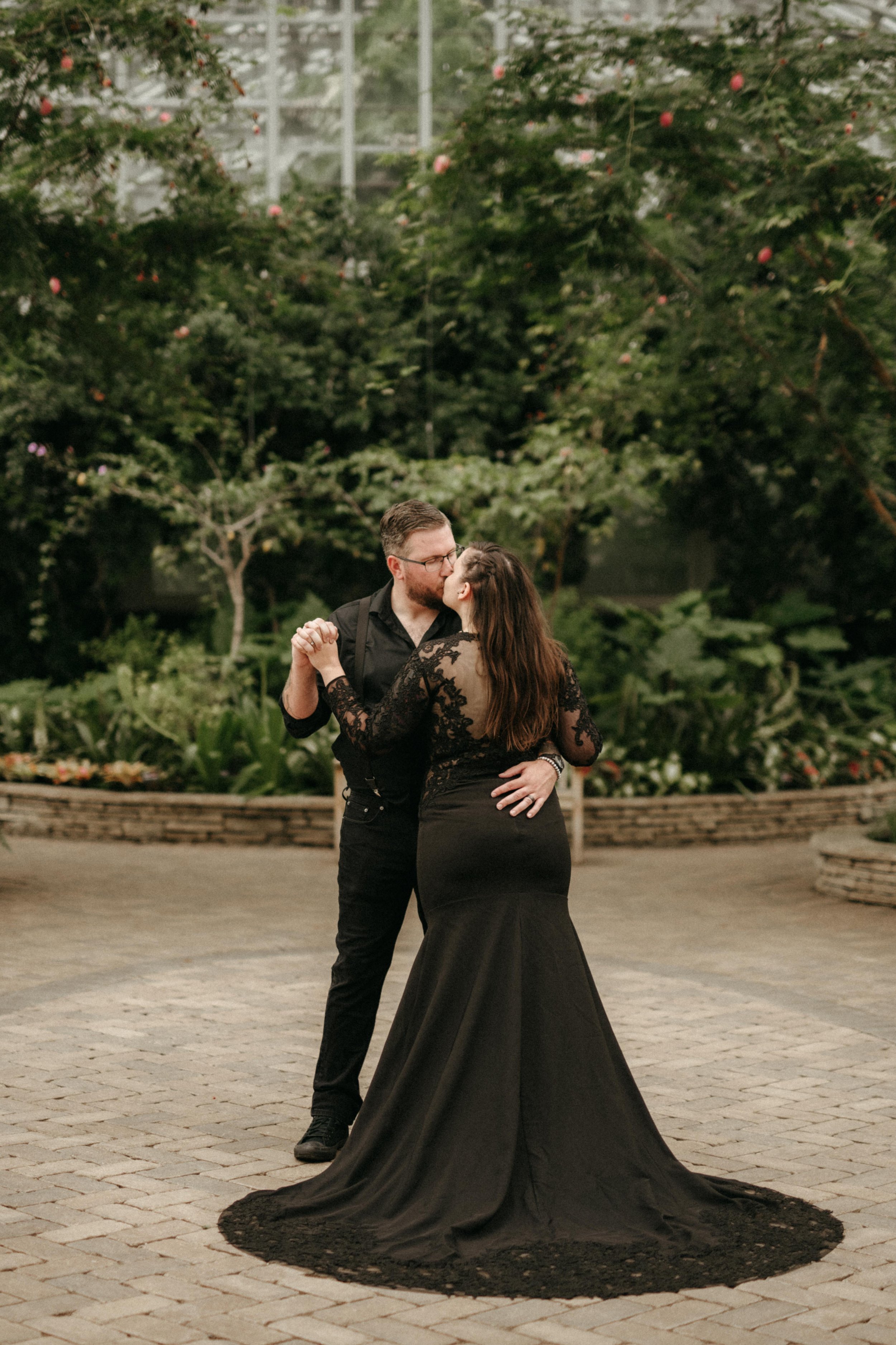Bride and groom in all black kiss in a greenhouse with tall green trees and a glass wall.