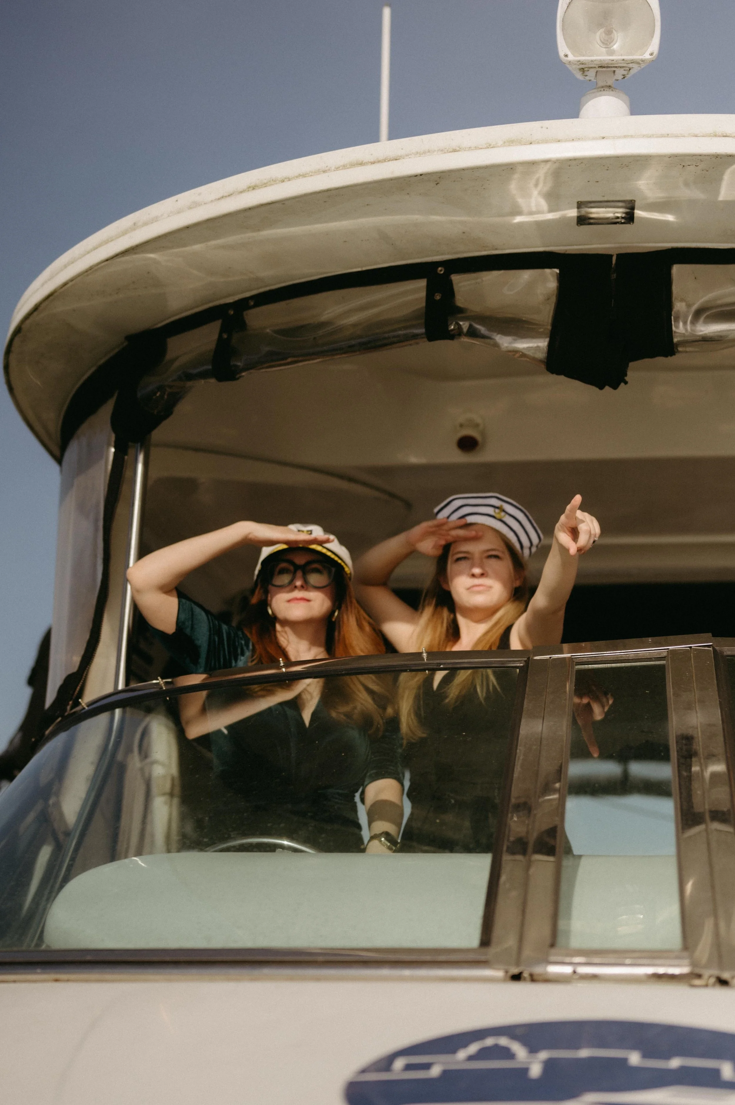 Wedding guests wearing captains hats and pointing into the distance on a dinner cruise rehearsal dinner on Lake Mendota. Moody Madison wedding photographer.