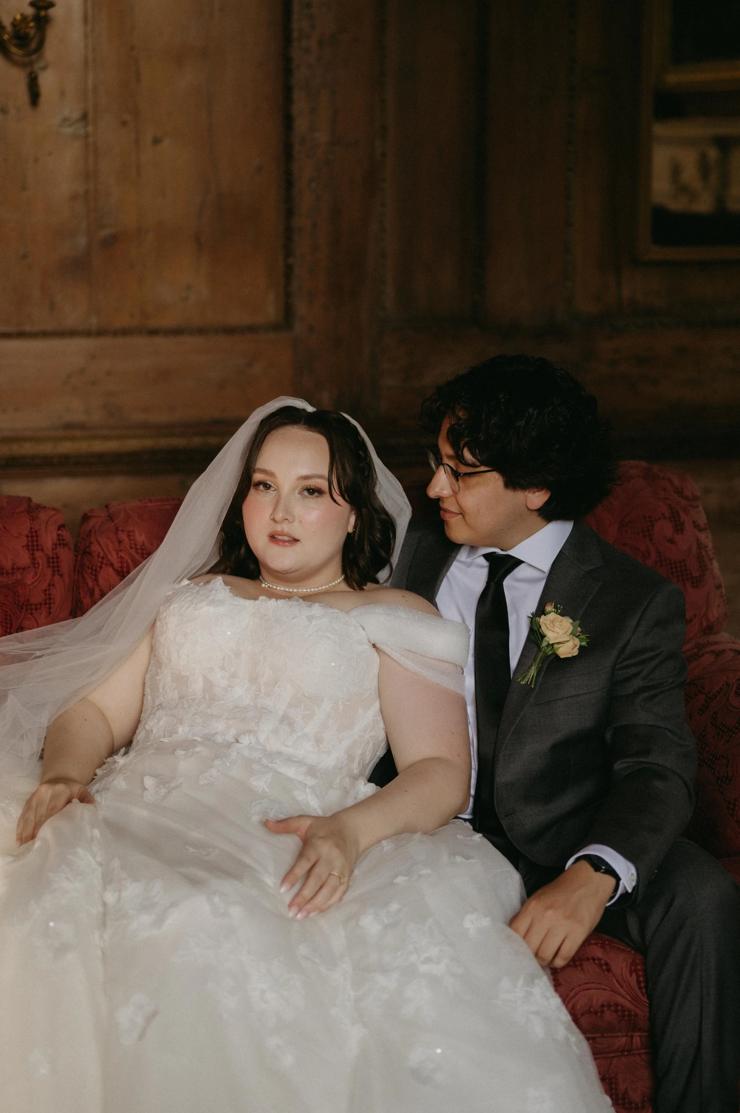 A bride and groom lounge on a red couch inside Cedar Hall in Memphis, Tennessee. The bride has short hair, a long white veil, and a lacey white wedding dress with off the shoulder sleeves. The groom wears a dark grey suit with a yellow boutonniere.
