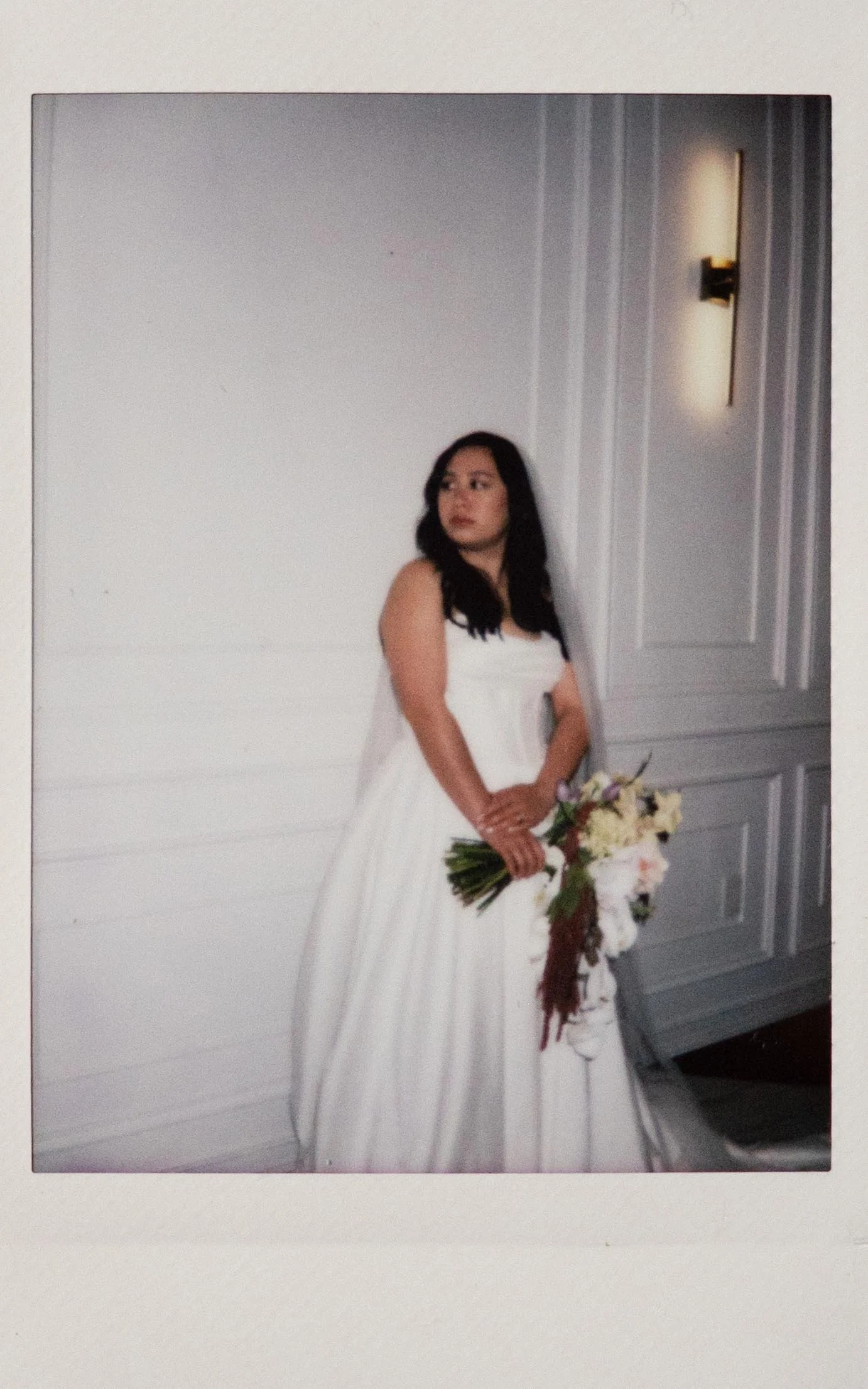 Moody film bridal portait on polaroid instant film, a bride in a white ball gown poses against a white wall in the Parisian in Huntsville, Alabama. She holds a yellow and white bouquet.