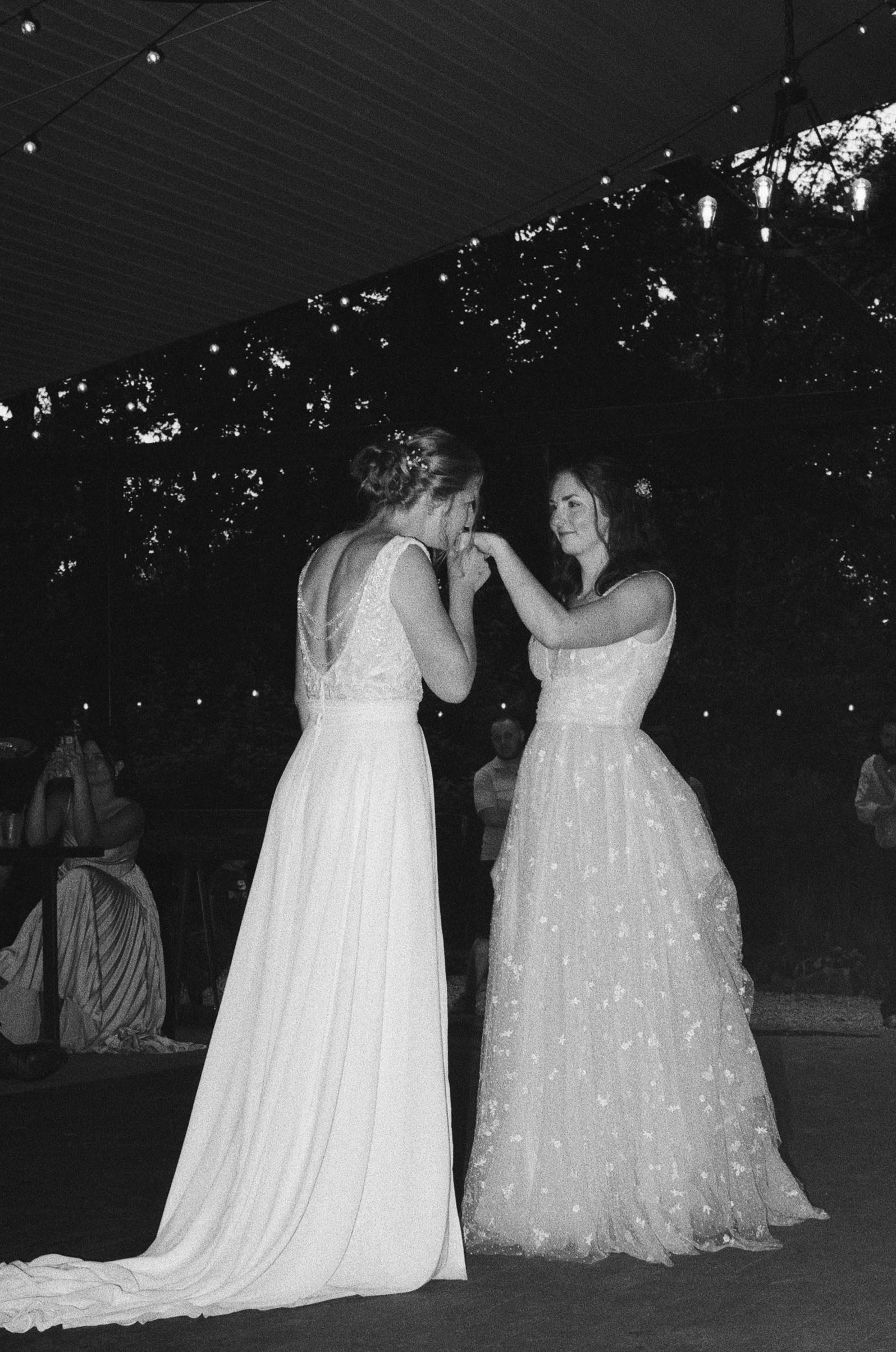 Black and white 35mm photo of two brides beginning their first dance. Once bride kisses the other's hand. One dress is tulle with embroidered flowers. The other bride wears a jumpsuit with a skirt attachment.
