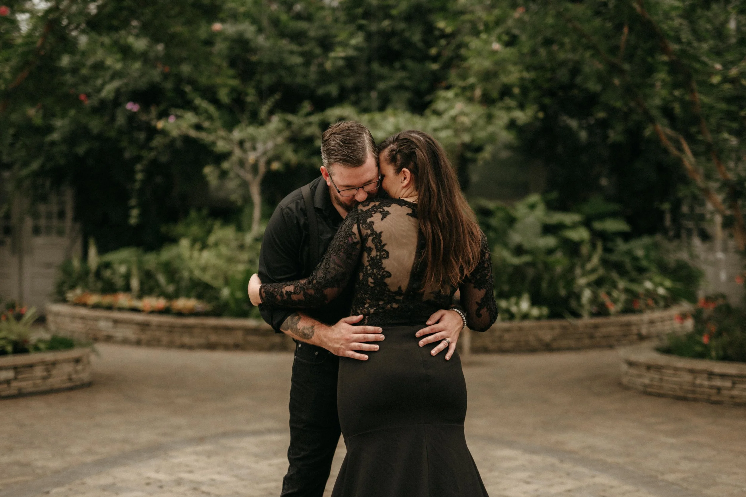 Editorial couple pose with the groom kissing the bride's shoulder. They are wearing all black and posing in front of greenery in a greenhouse.