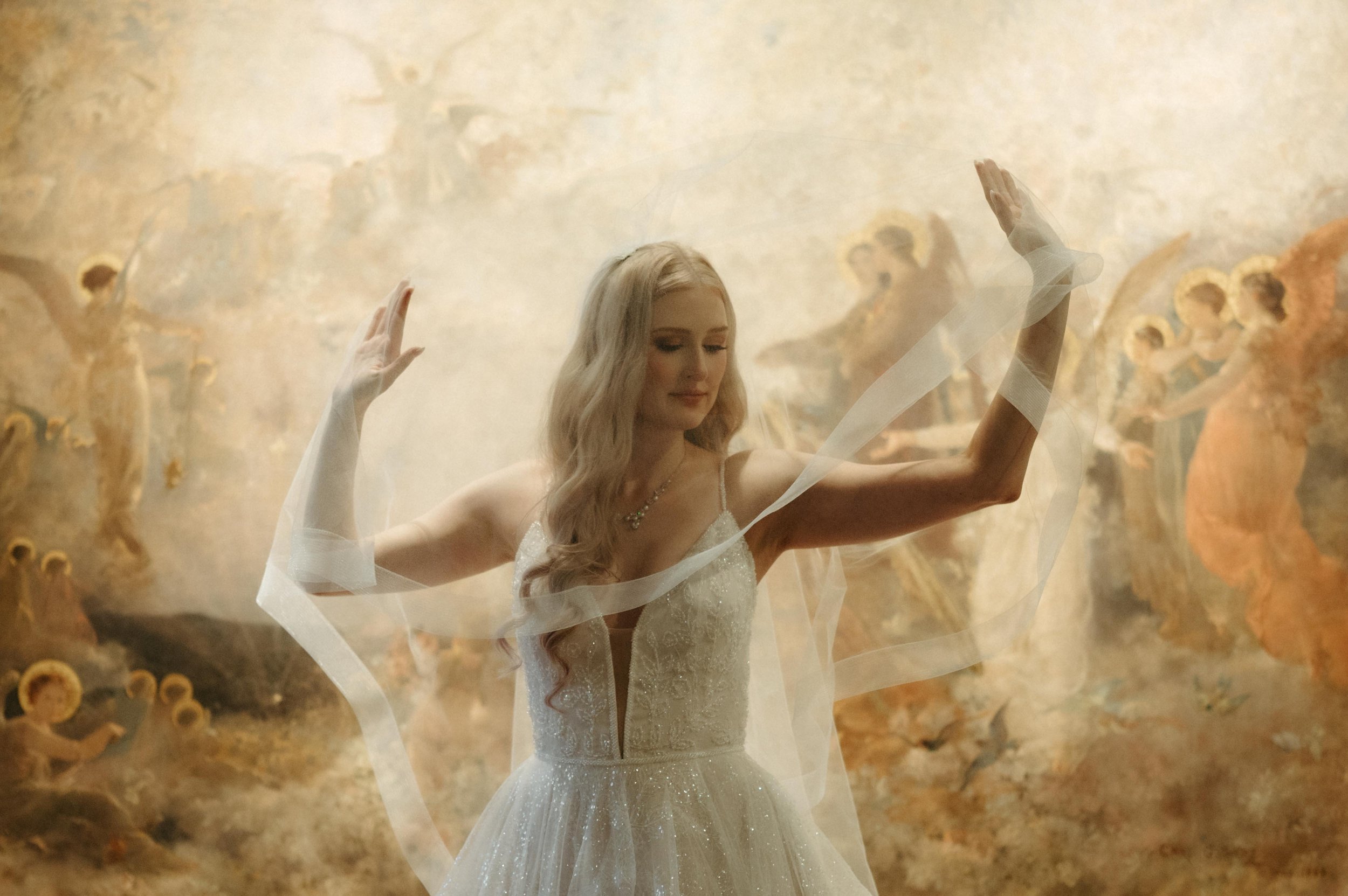 A bride stands in front of a golden hued painting at the Brooks Museum of Art in Memphis, Tennessee, lifting her veil over her head in a dramatic, editorial bridal pose. She wears her long blonde hair down over one shoulder, and a sparkly, thin-strap