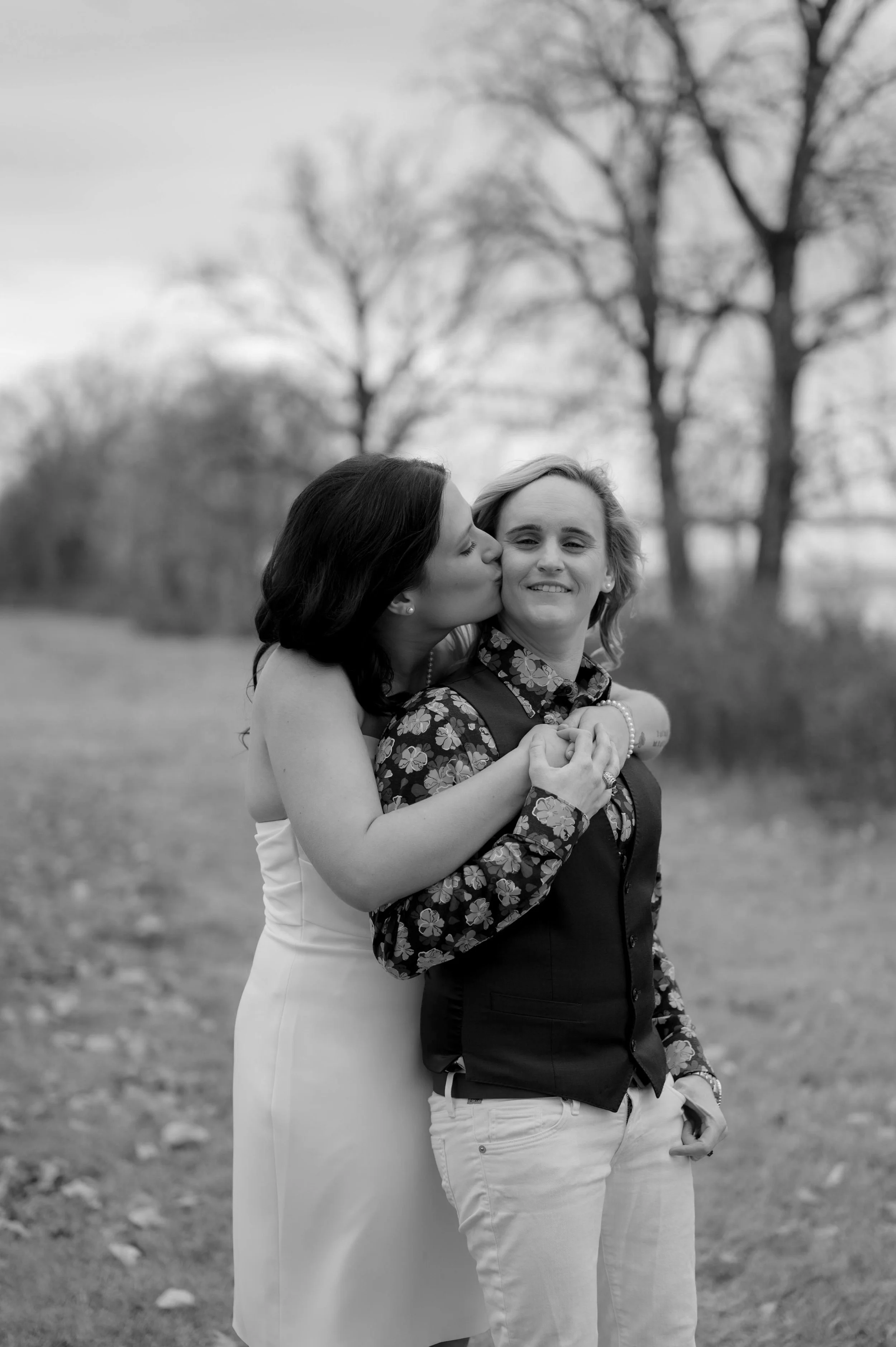 Two brides pose in front of the riverfront in downtown Memphis, Tennessee. One bride wears a white mini dress and embraces the second bride who wears a floral top and a vest.
