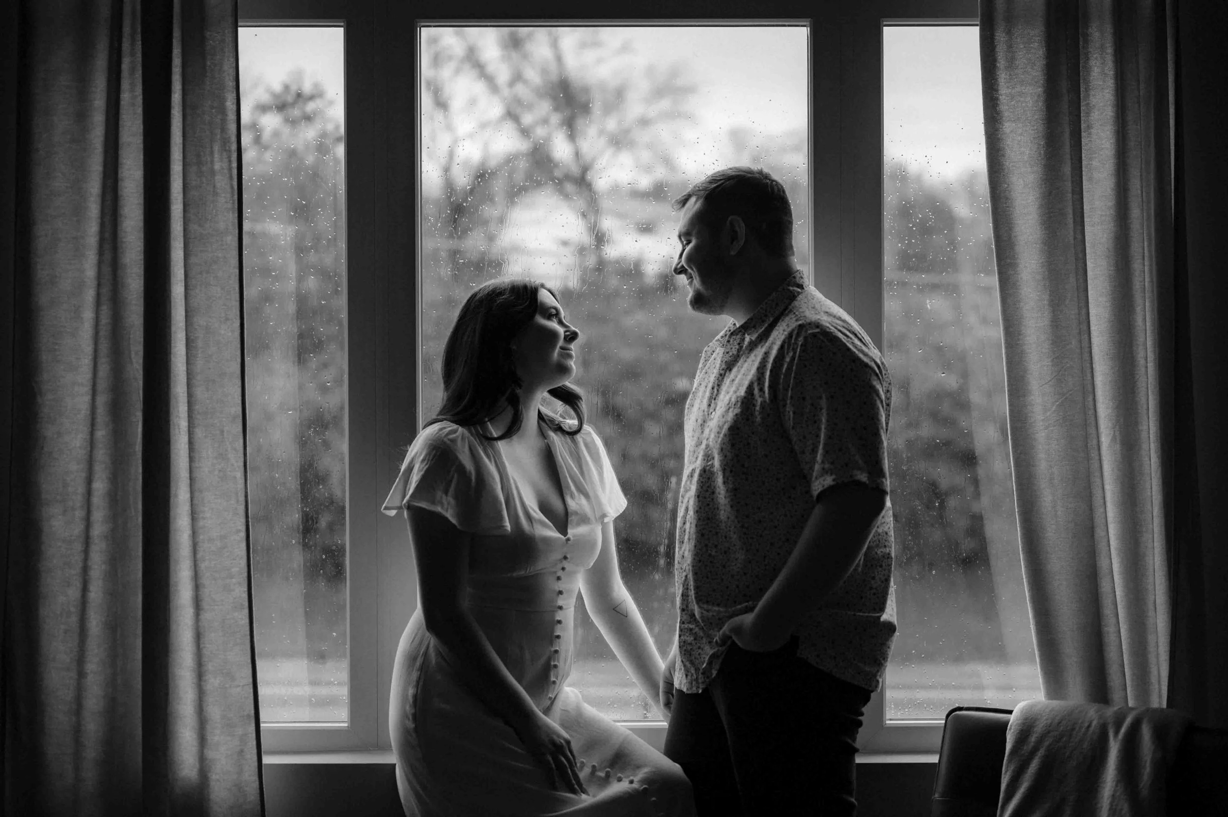 Moody black and white photo of a couple posing in front of a rainy window in Nashville, Tennessee. Moody editorial Nashville wedding photographer.