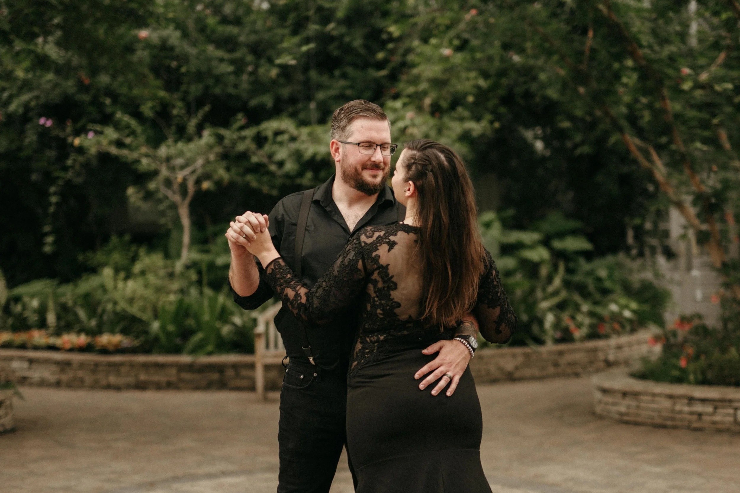 A couple in all black face each other, holding hans. They wear all black and pose inside the Garfield Park Conservatory in Chicago, Illinois.