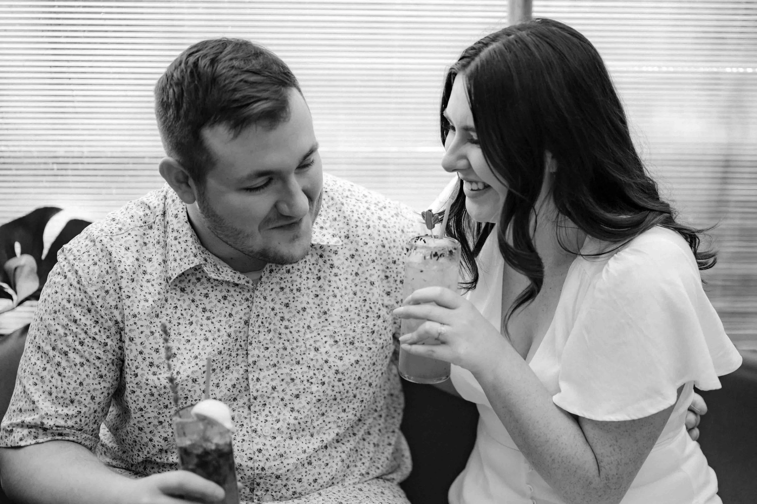 Black and white photo of a couple dressed in white and drink cocktails, laughing and smiling at each other.