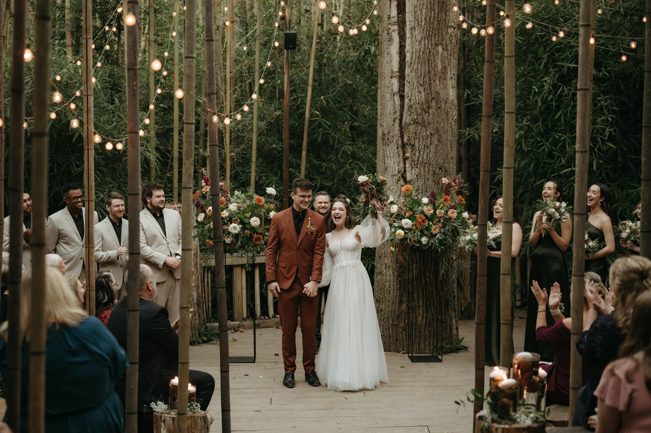 A couple celebrates at the end of their ceremony in the bamboo forest at Juniper Farms outside Huntsville Alabama. The groom wears a terracotta orange suit, and the groomsmen wear tan suits. The bride wears a white dress with poet sleeves, and the br