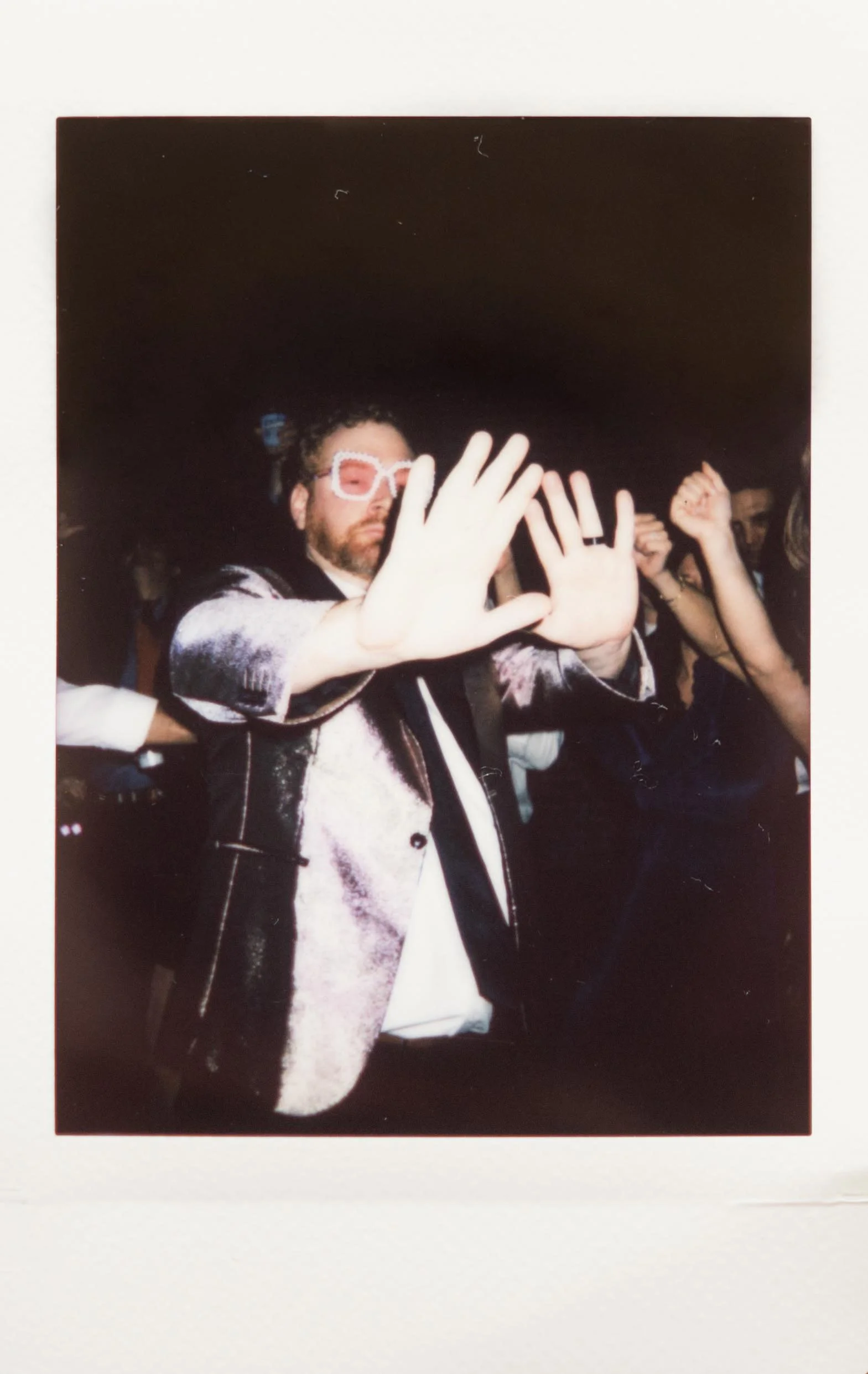 Polaroid photo of a groom dancing at The Kent in Memphis, Tennessee. He has his hands out towards the camera, and he's wearing a shiny purple suit jacket and white and pink sunglasses. Other guests have their hands up behind him.