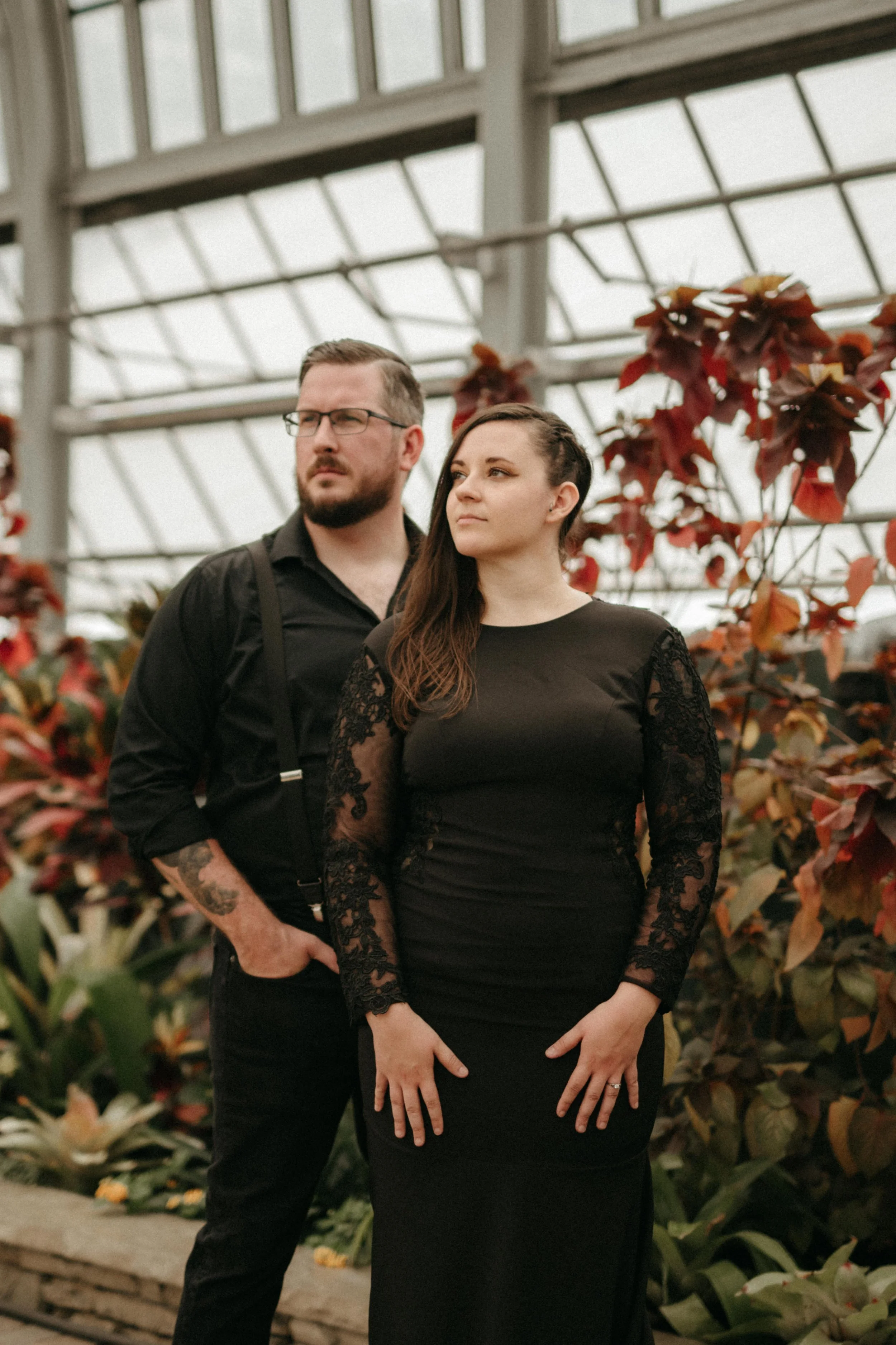 A couple in all black wedding attire stands in an editorial bridal pose in front of orange and red plants in the Garfield Park Conservatory in Chicago, Illinois.