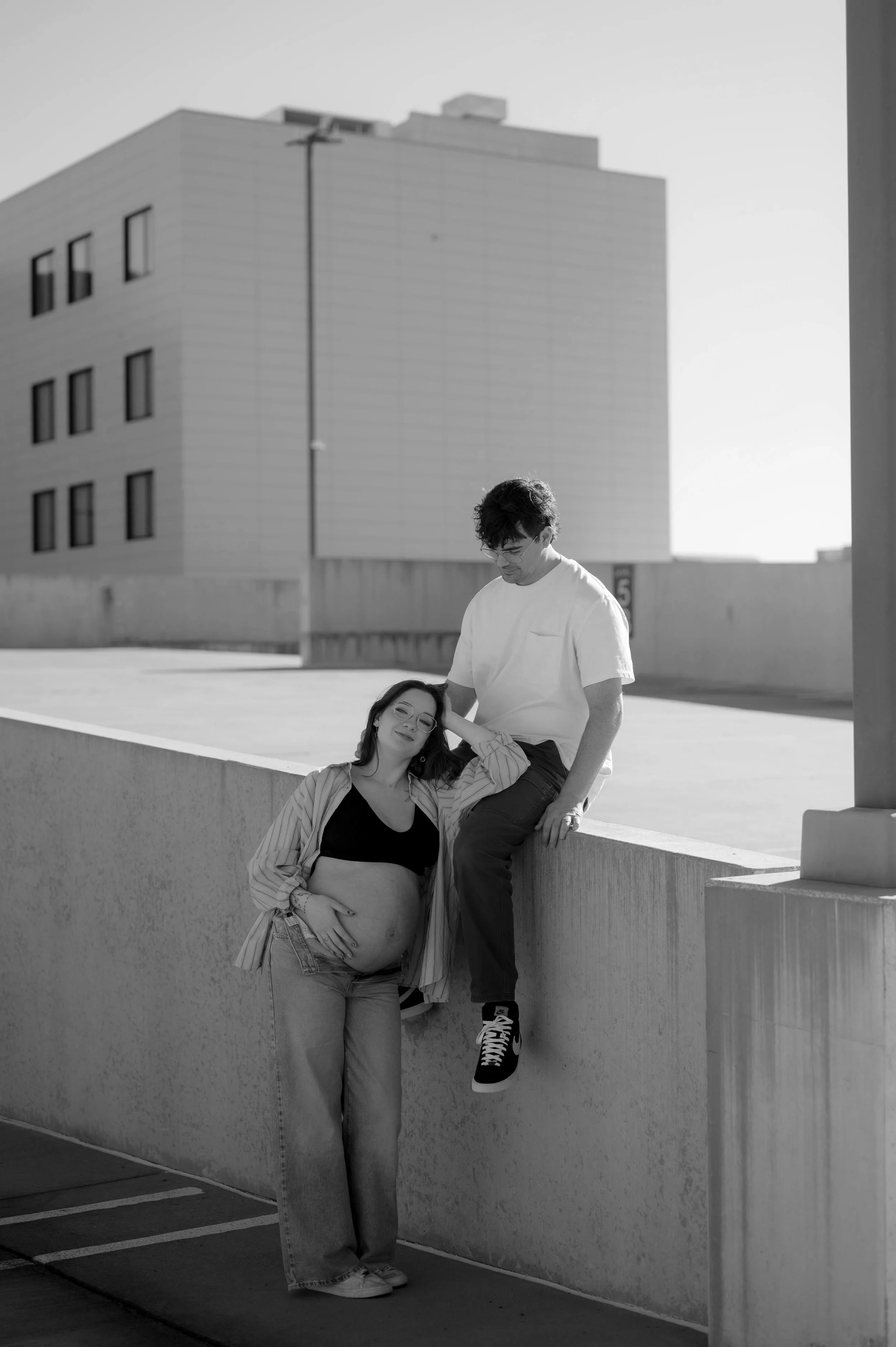 Black and white editorial maternity session on a rooftop in downtown Huntsville, Alabama. The mom wears denim and an open button down to show off her baby bump, and the dad sits on a wall above her.