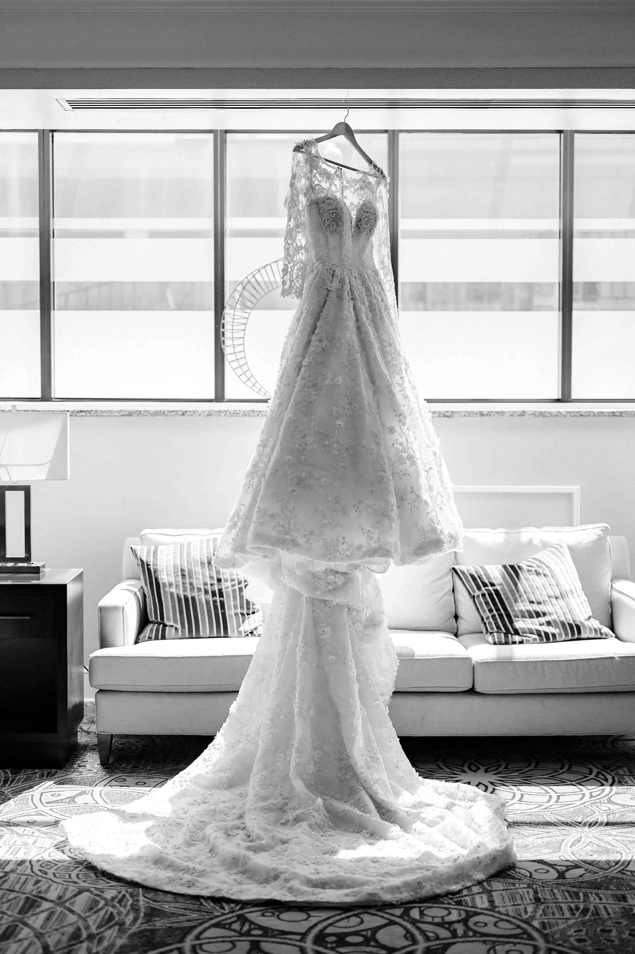 Black and white photo of a beaded wedding dress hanging in front of a window in the Peabody Hotel in Memphis, Tennessee. The dress has intricate beading, long sheer lace sleeves and a dramatic train.