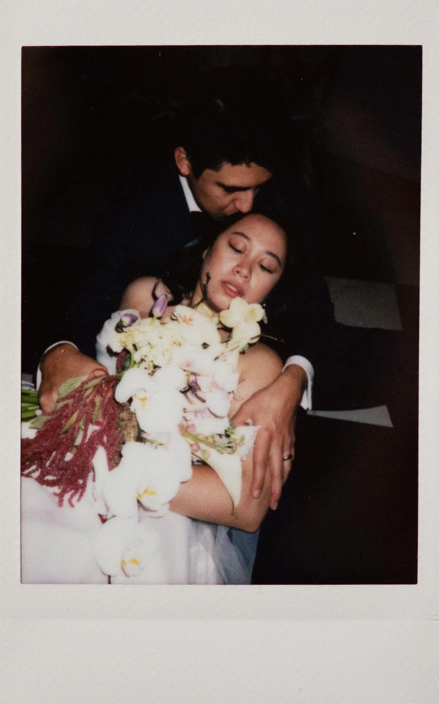 Polaroid photo of a couple in The Parisian in Huntsville, Alabama. The bride is laying across the groom's lap in an editorial pose, holding a large bouquet of while and red flowers. The groom kisses her forhead.