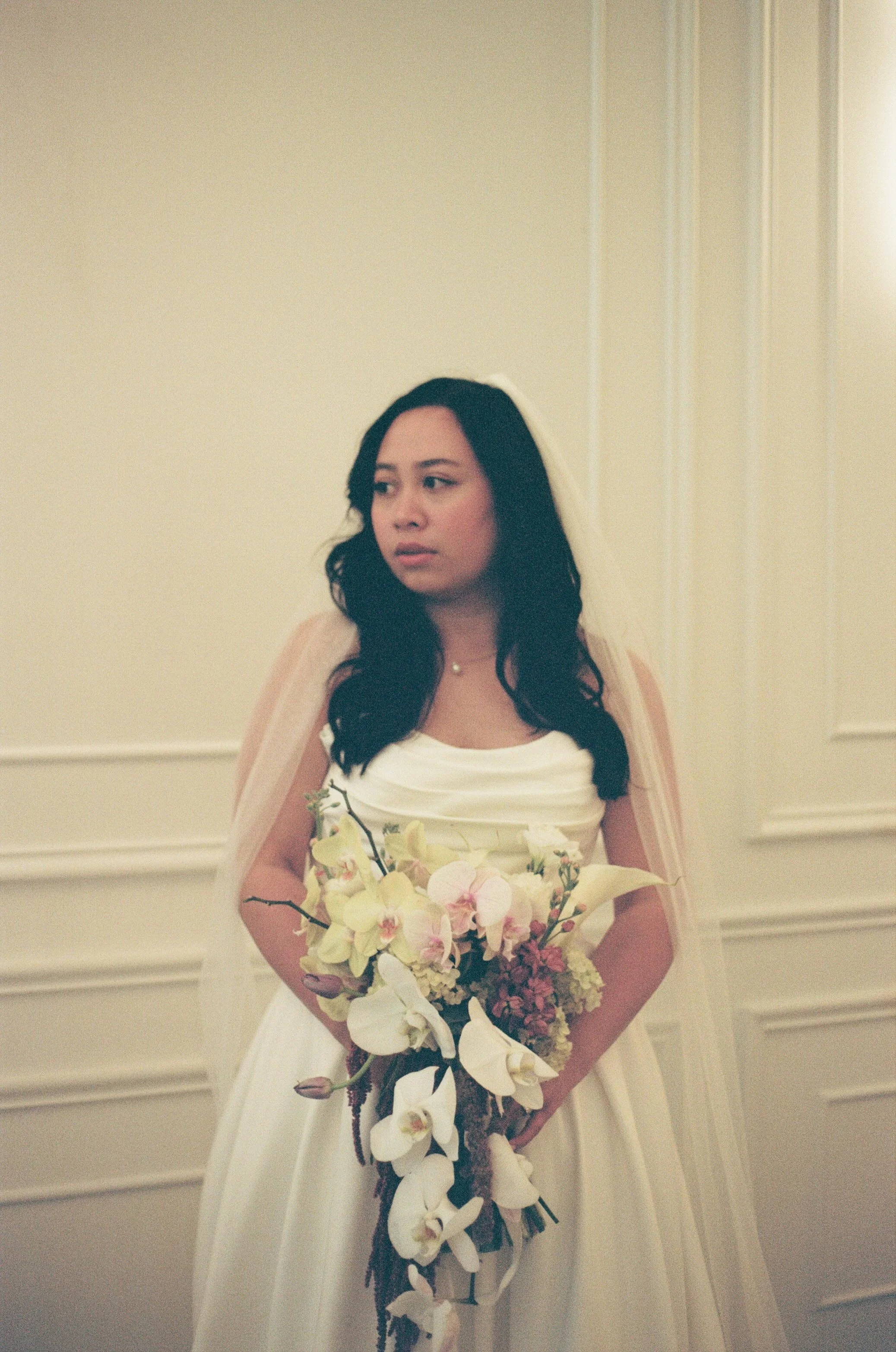 A 35mm photo of a bride posing in front of a white wall in The Parisian in Huntsville, Alabama. She wears a classic simple white gown and holds a large white, pink, and yellow bouquet made by Noella's Flowers in Huntsville.