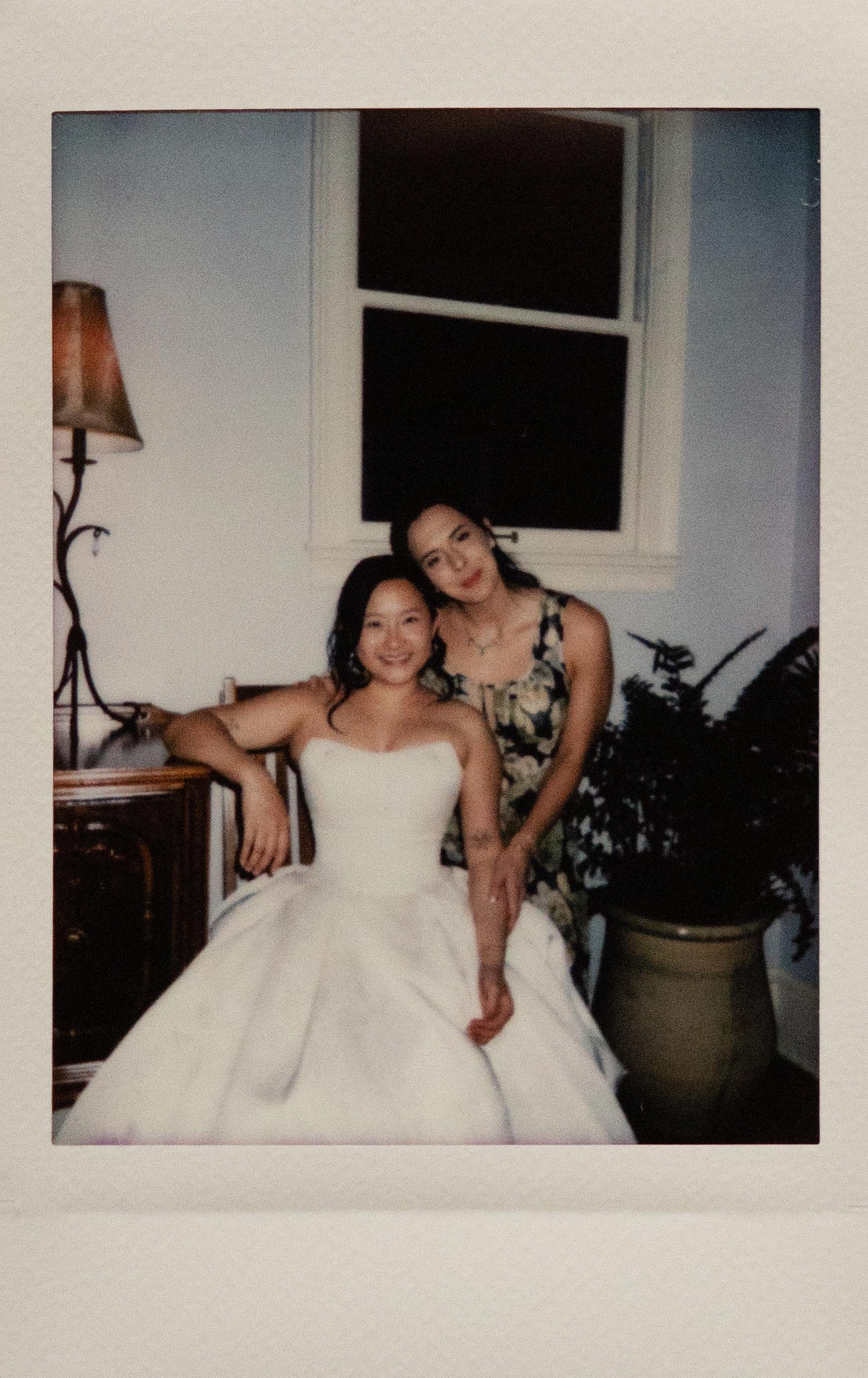 Polaroid photo of a bride seated, wear a simple strapless white gown with a ballgown skirt. A bridesmaid in a floral pattern dress leans over her. They are in a home in Midtown Memphis, Tennessee.