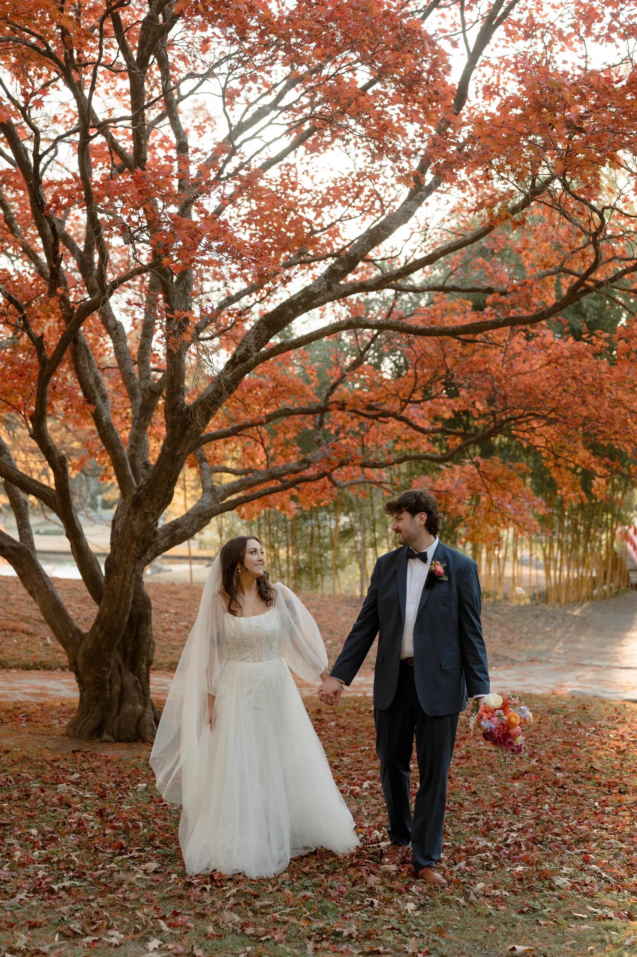 A bride and groom walk under an orange maple tree at the Memphis Botanic Garden in Memphis, Tennessee. The bride wears a long sleeve white wedding dress with a tulle skirt, and the groom wears a blue suit.