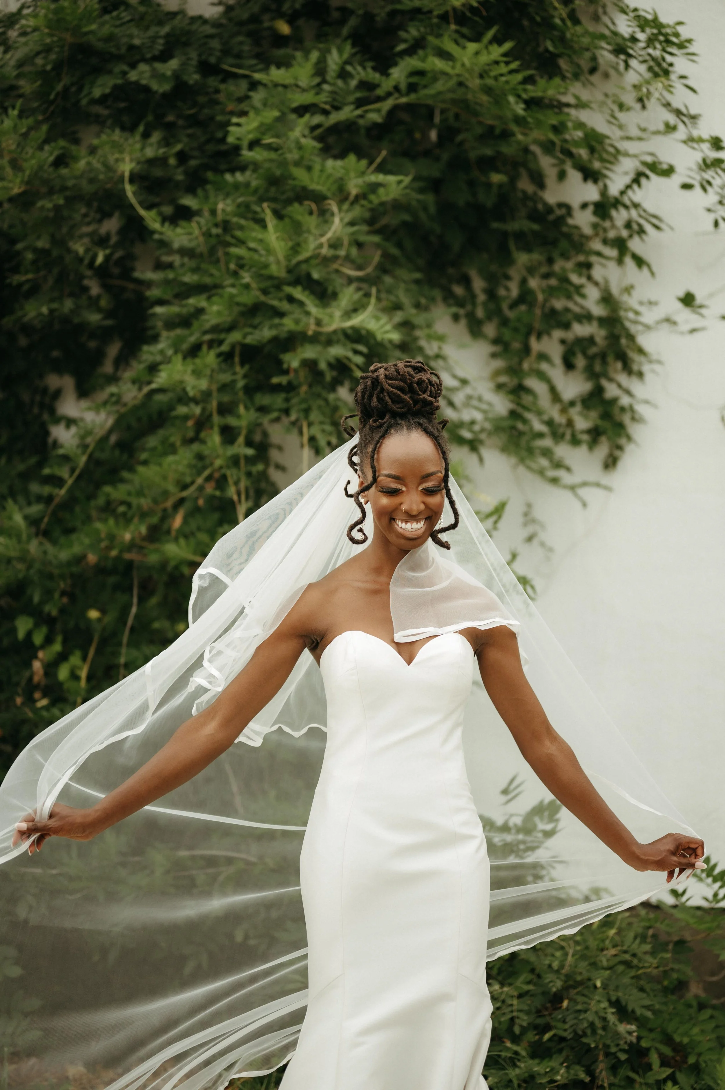 Editorial wedding photography of a bride wearing a white strapless gown and a long white veil that blows in the wind. She poses in front of greenery at the Plant Venue in Jackson, Mississippi.