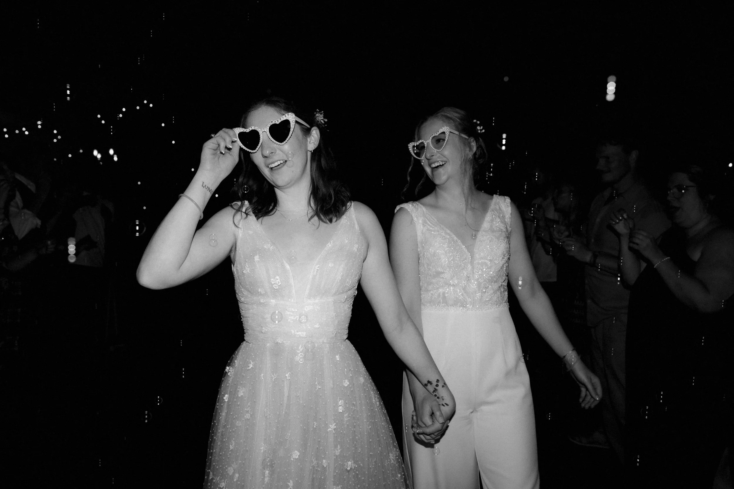 Black and white photo of two brides running through a bubble exit at the end of their reception. They're holding hands and wearing white heart shaped sunglasses.