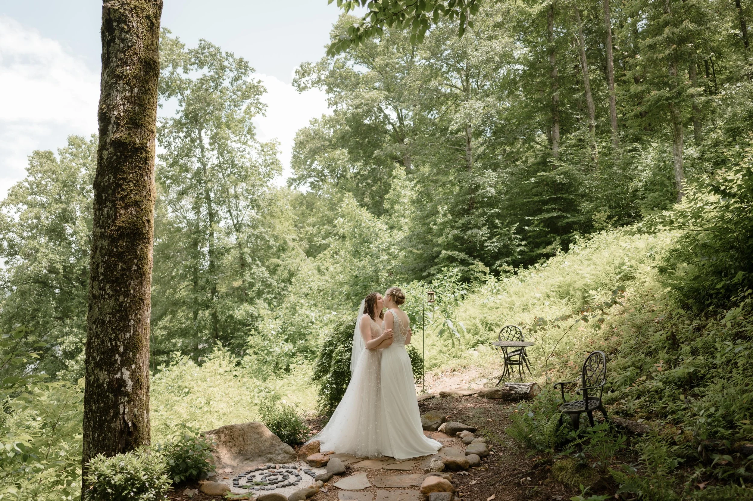 Two brides share a kiss during their first look on a mountainside in Nantahala, North Carolina. One bride wears an off white dress with tulle and a veil, and the other bride wears a white jumpsuit with a long white skirt attachment.