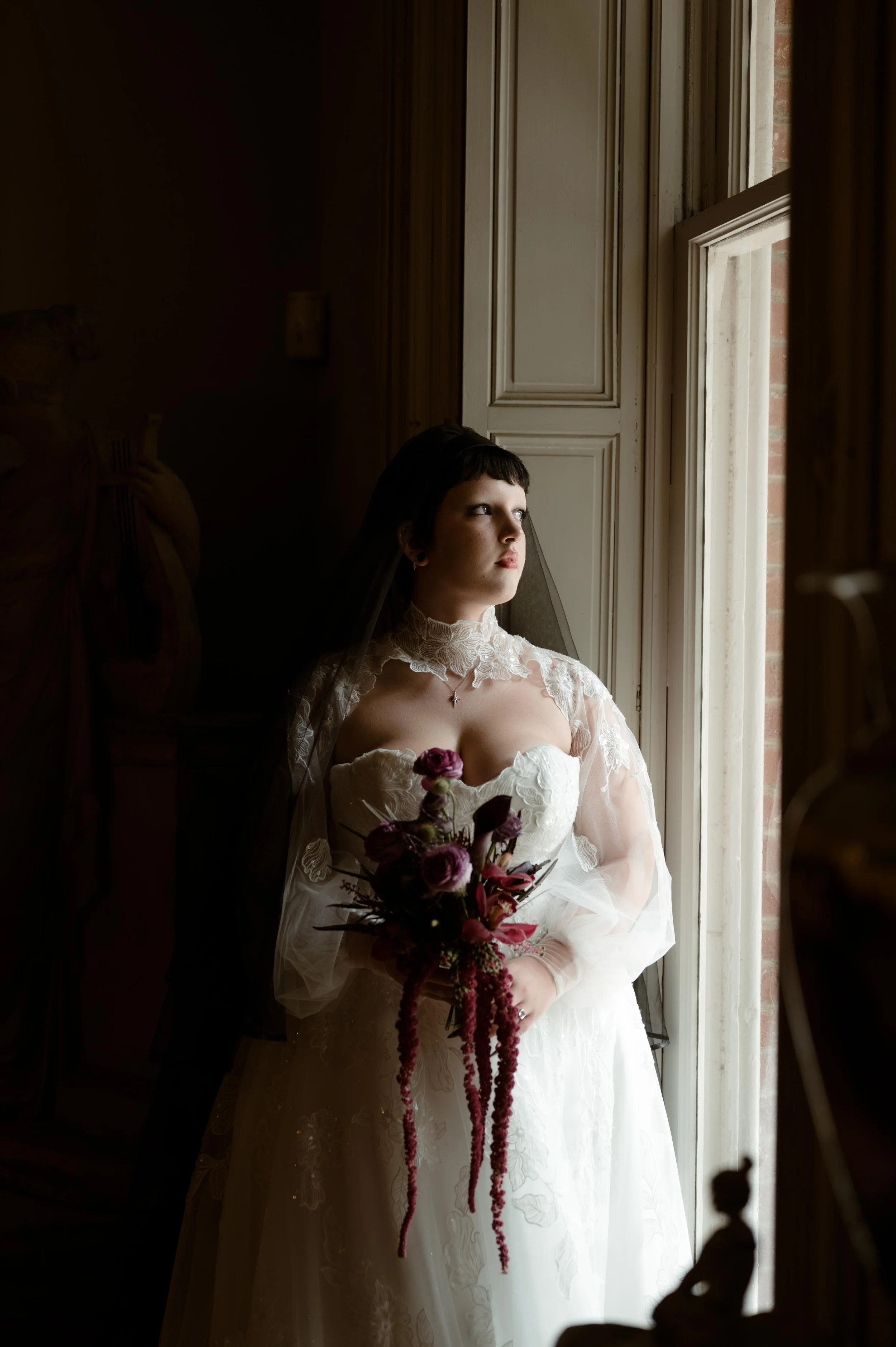 Moody wedding photography of a bride posting by a window in the Woodruff Fontaine House in Memphis, Tennessee. She holds a purple and red bouquet and wears a long black veil.